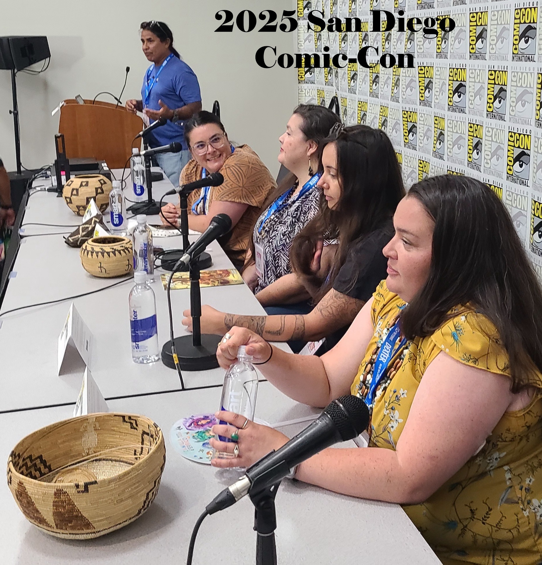 Panel of five women at a table during San Diego Comic-Con 2025, with a man standing behind them, microphones, and Comic-Con backdrop.