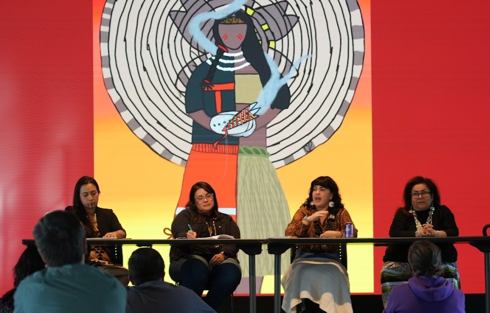 Four women sit at a panel table in front of a colorful mural of a person with a large circular background, speaking to an audience.
