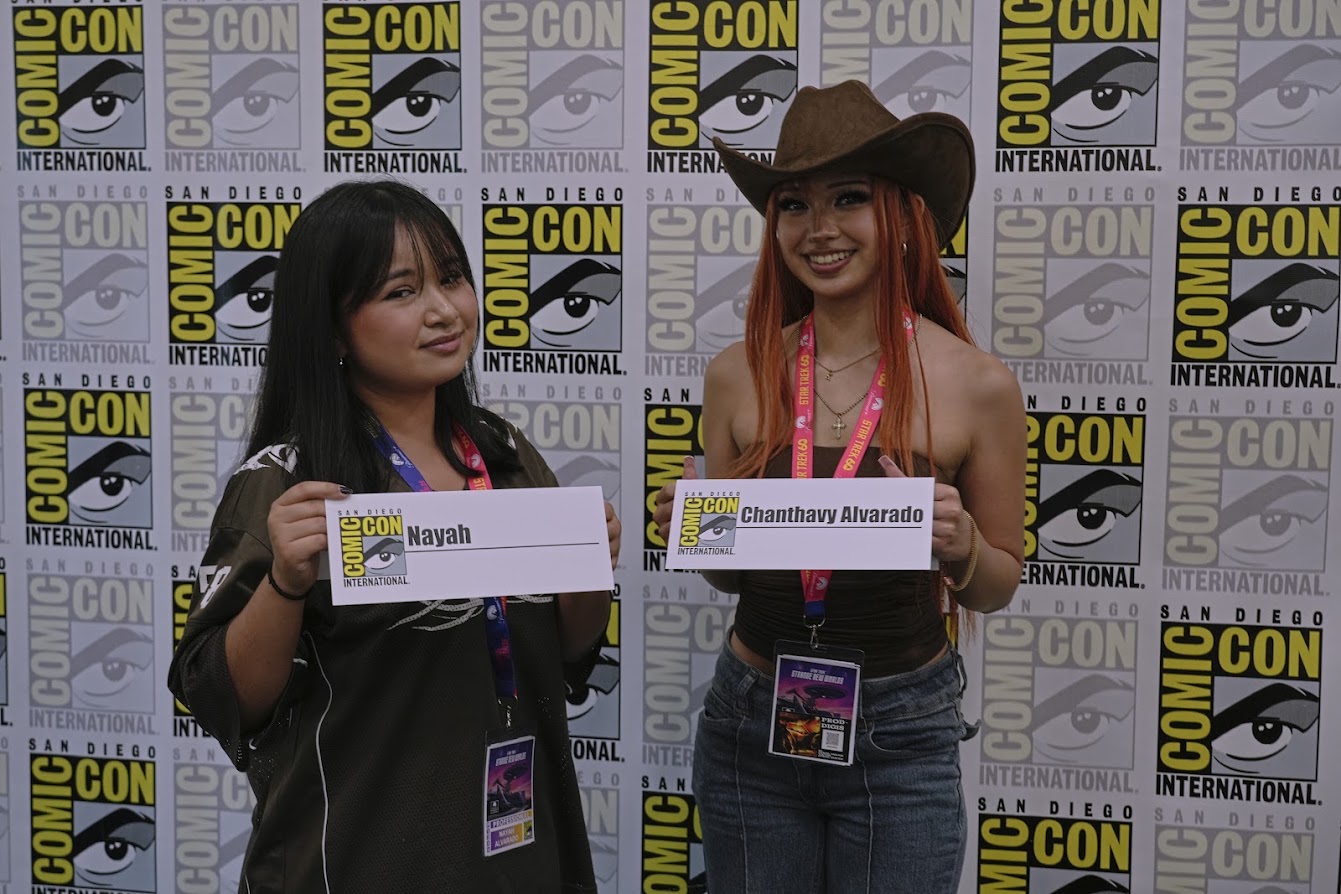 Two women holding signs in front of a Comic-Con International backdrop, one wearing a cowboy hat, both smiling.