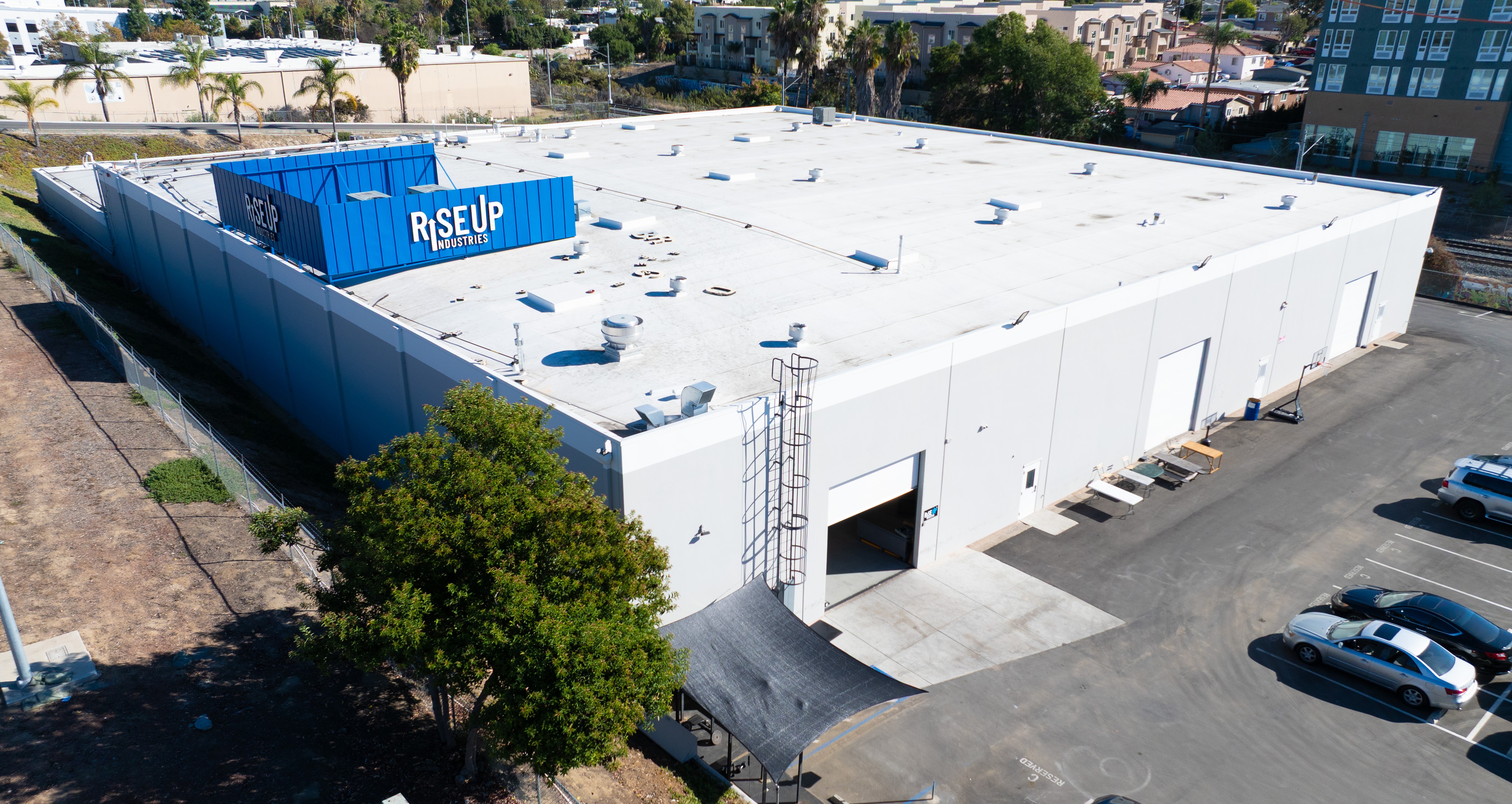 Large white industrial building with a blue sign reading 'RISE UP', surrounded by parking lot and trees.