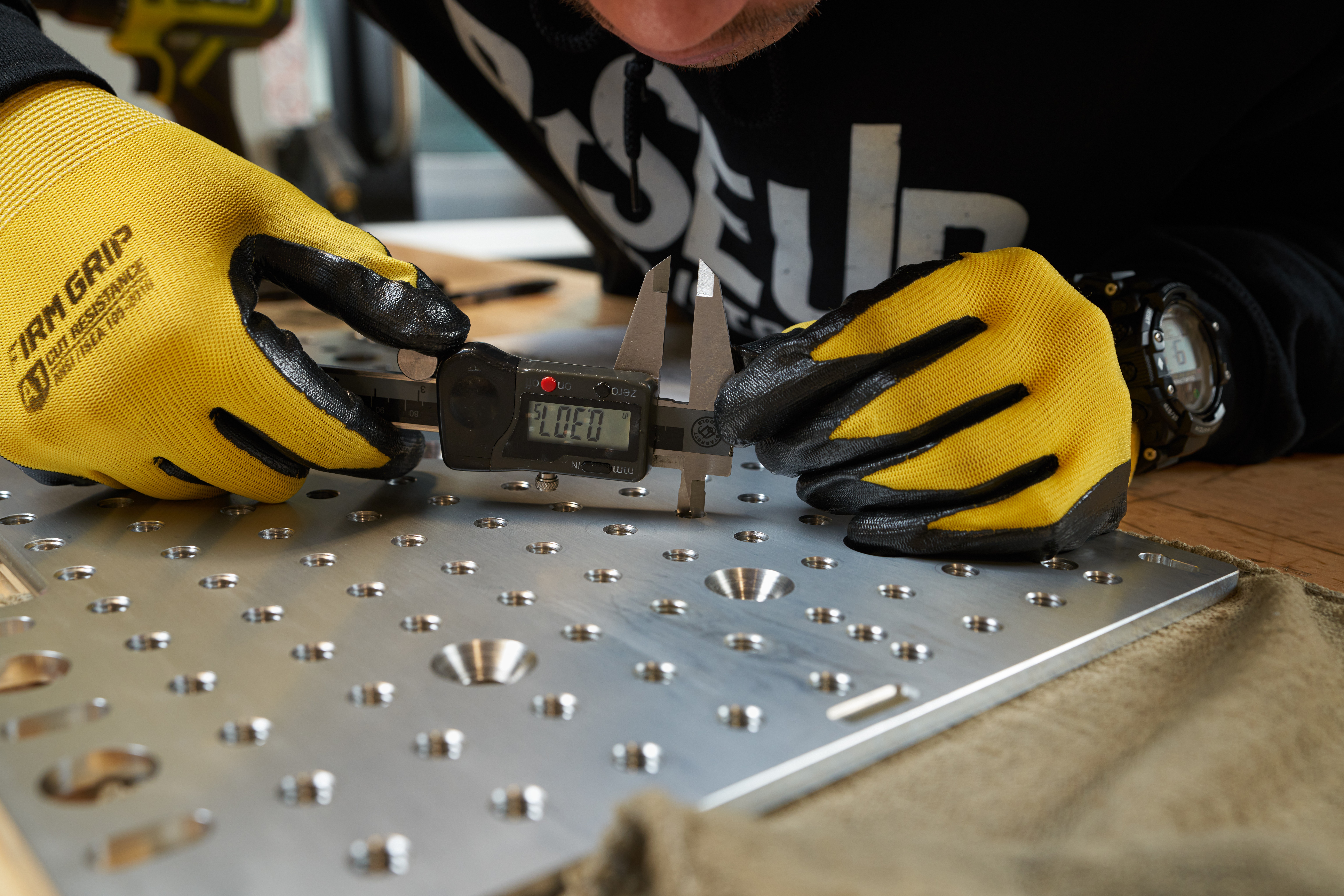 Person wearing yellow gloves using a digital caliper on a metal sheet with drilled holes, workspace visible in background.