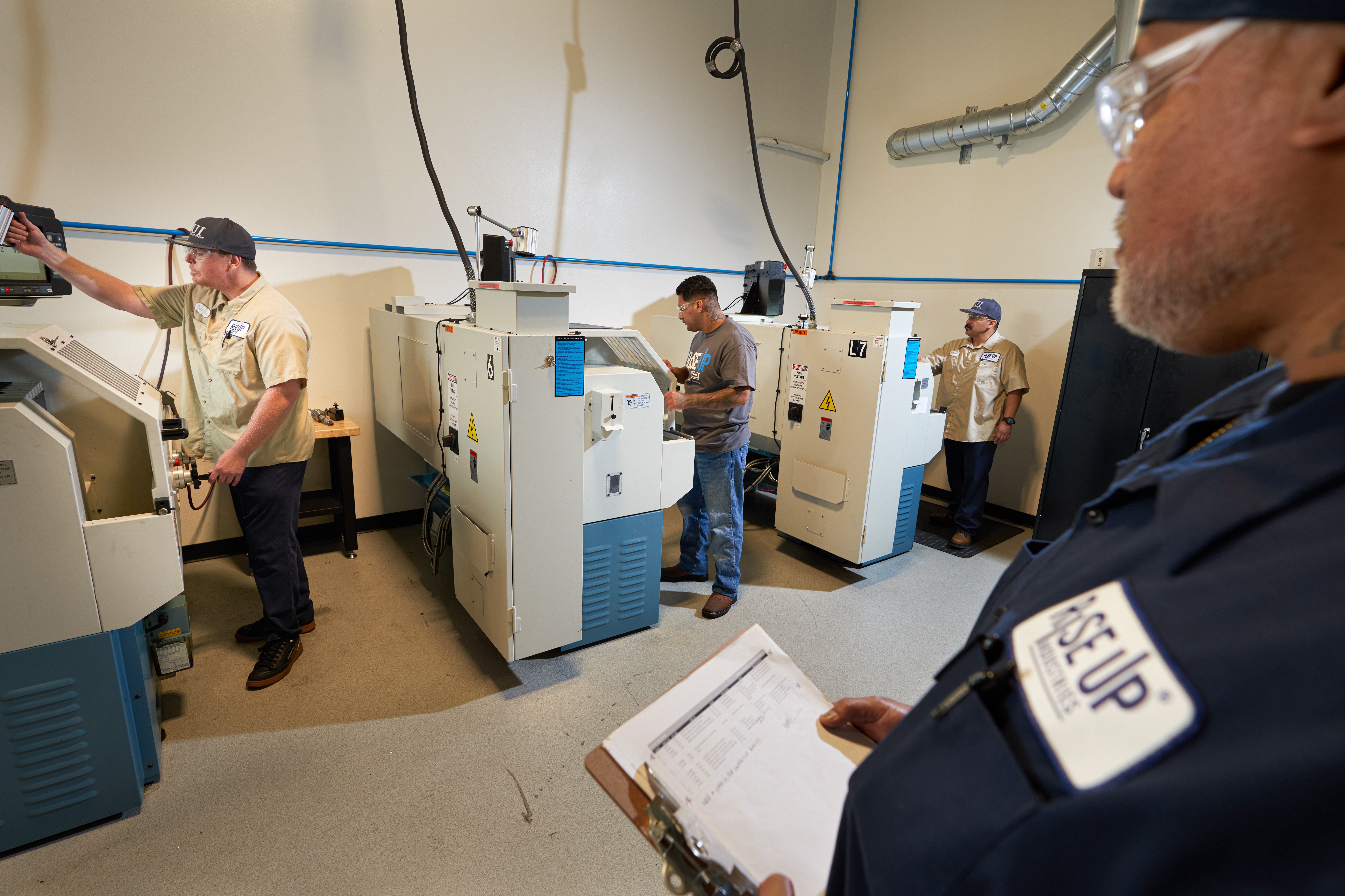 People working with machines in a laboratory or industrial setting, with a person in the foreground holding a clipboard.