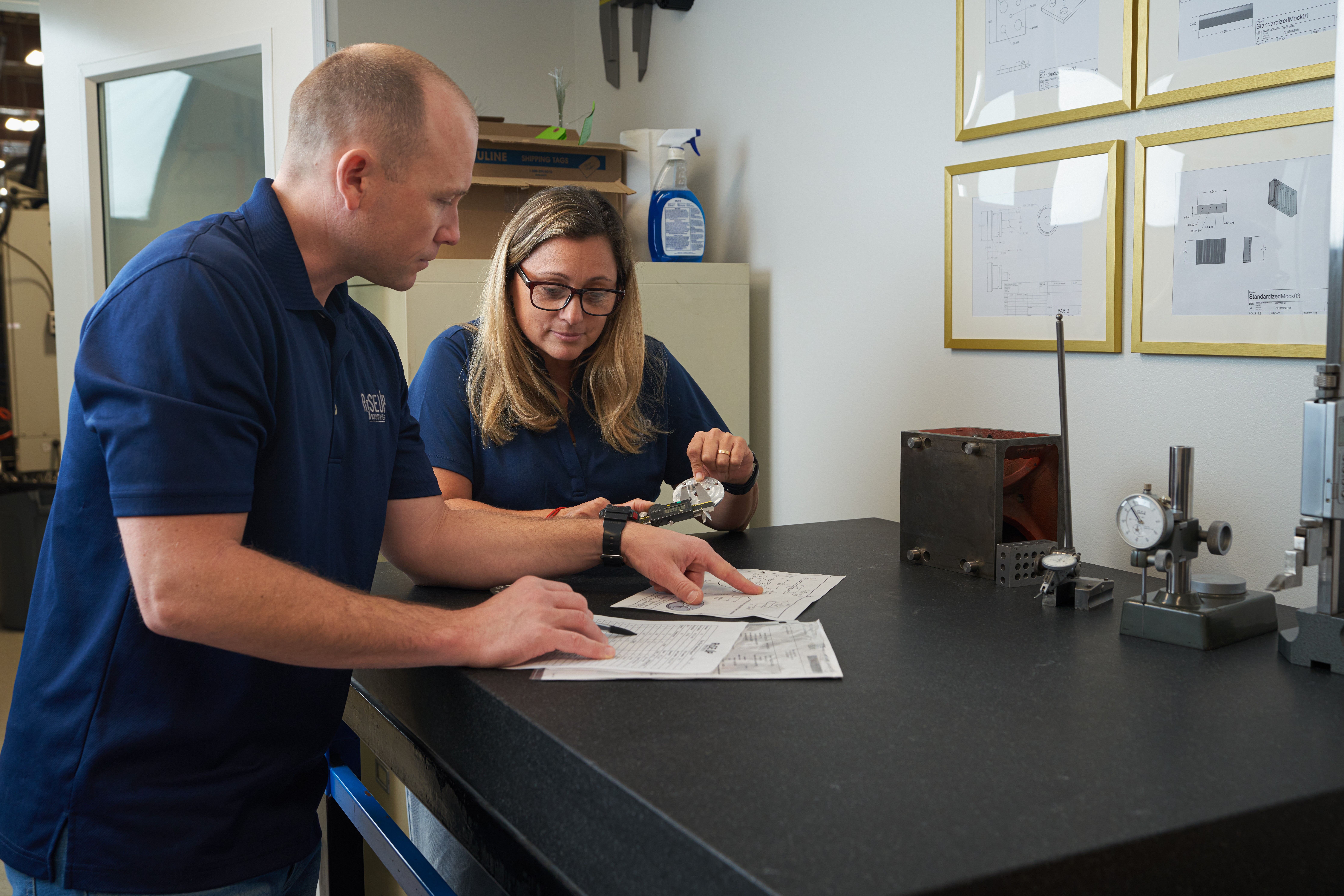 Two people in navy shirts review documents on a black table in a room with framed papers on the wall.