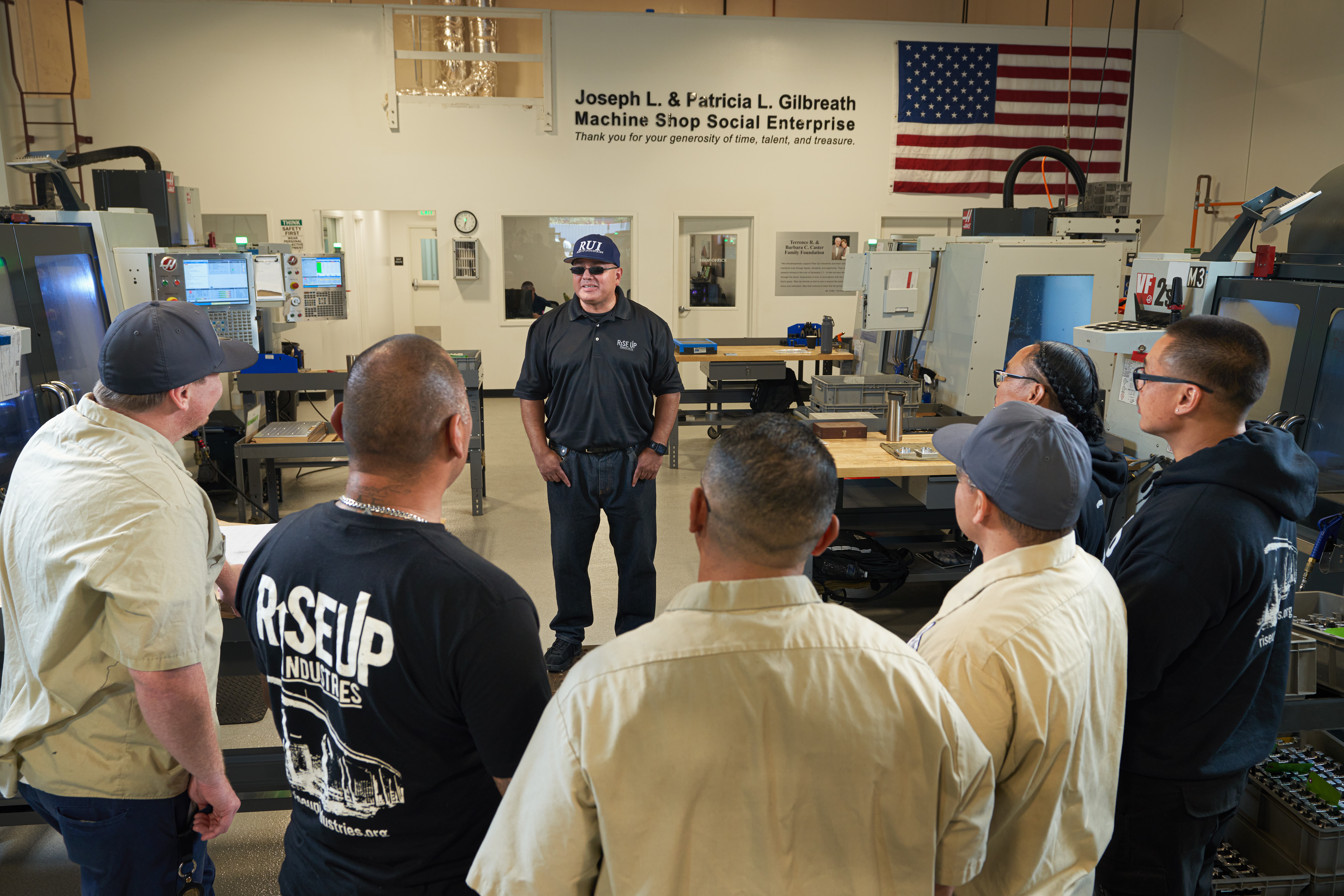 Group of people listening to a person in a machine shop with computers and an American flag in the background.