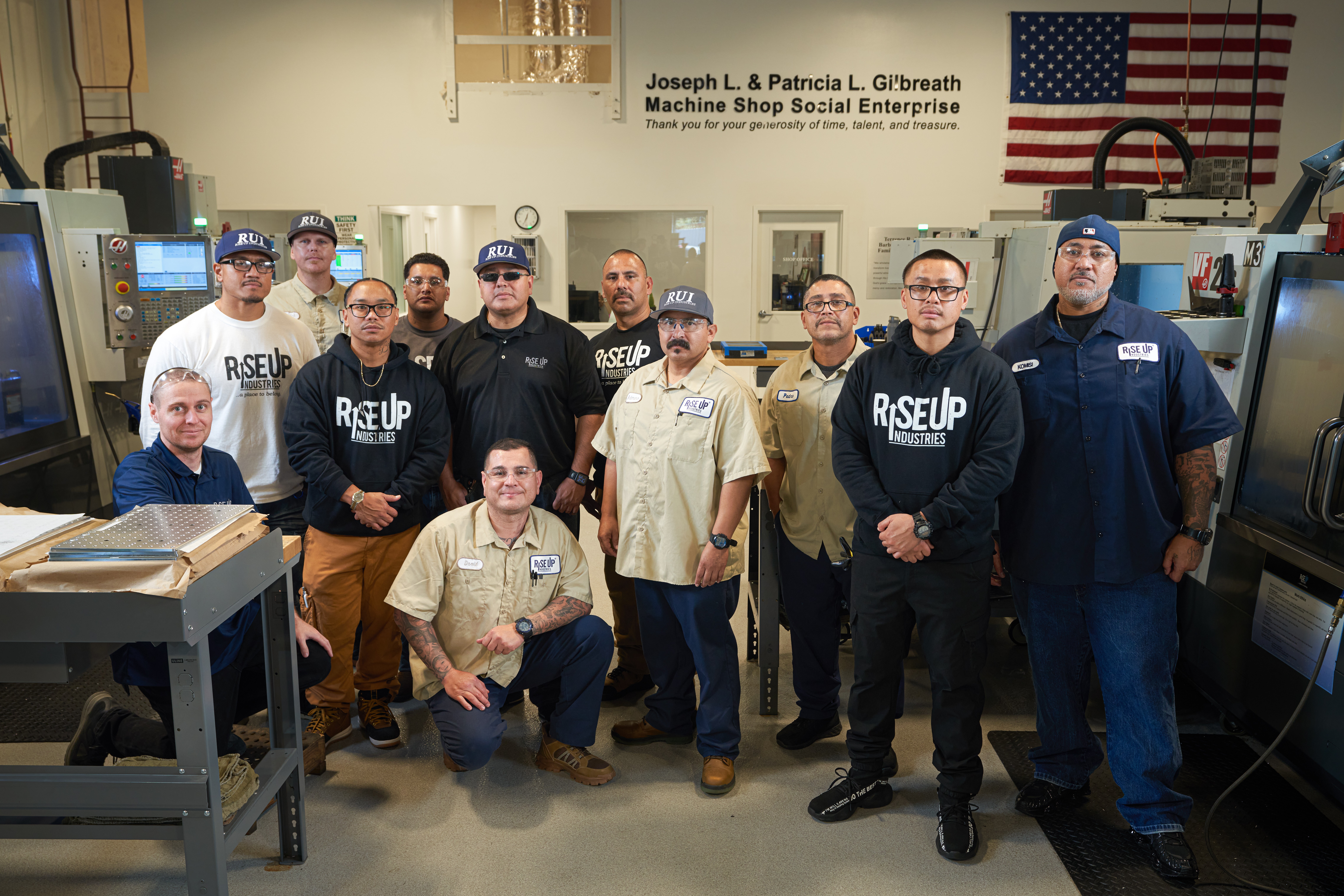Group of people in a workshop with machines and an American flag in the background, some wearing uniforms and others casual.