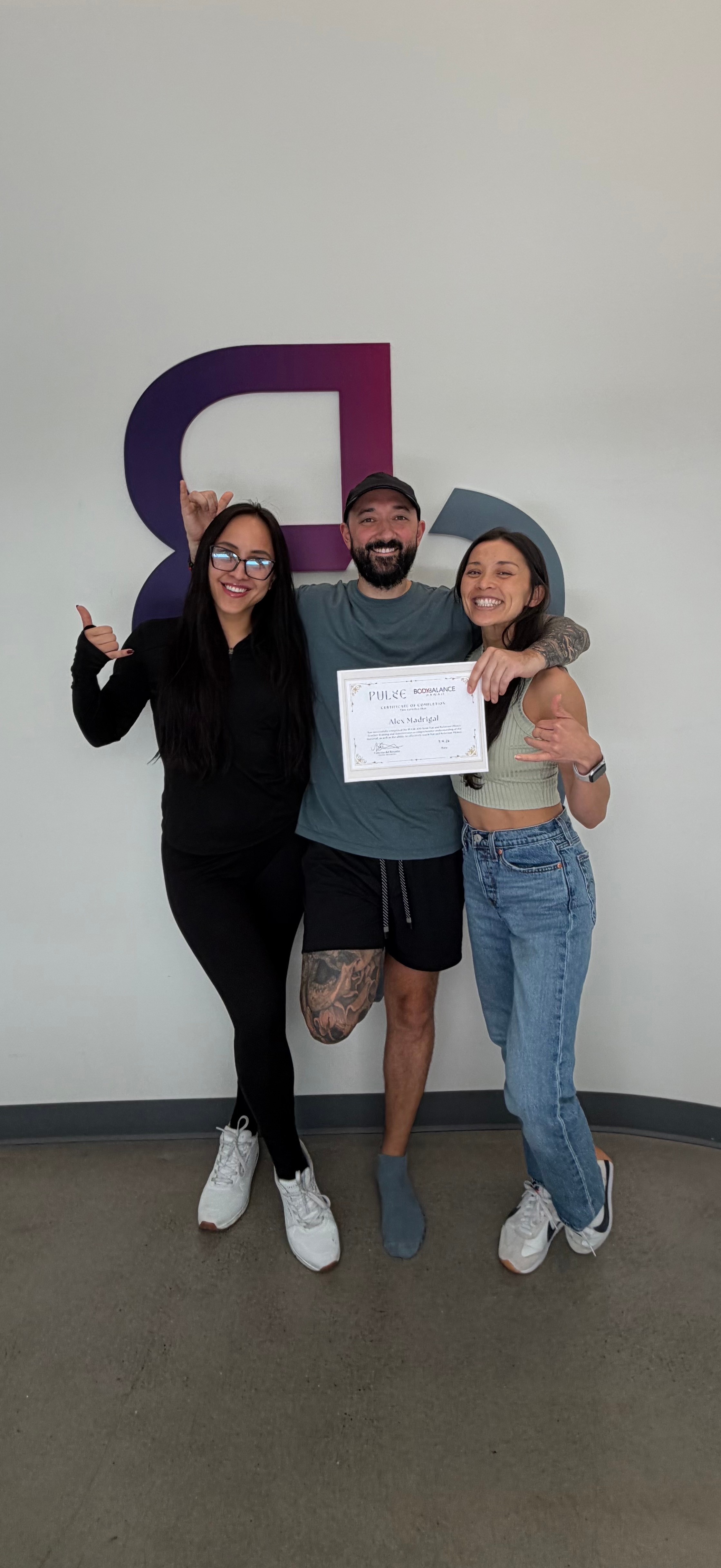 Three people standing together, one holding a certificate, smiling, in front of a wall with a large purple letter D.