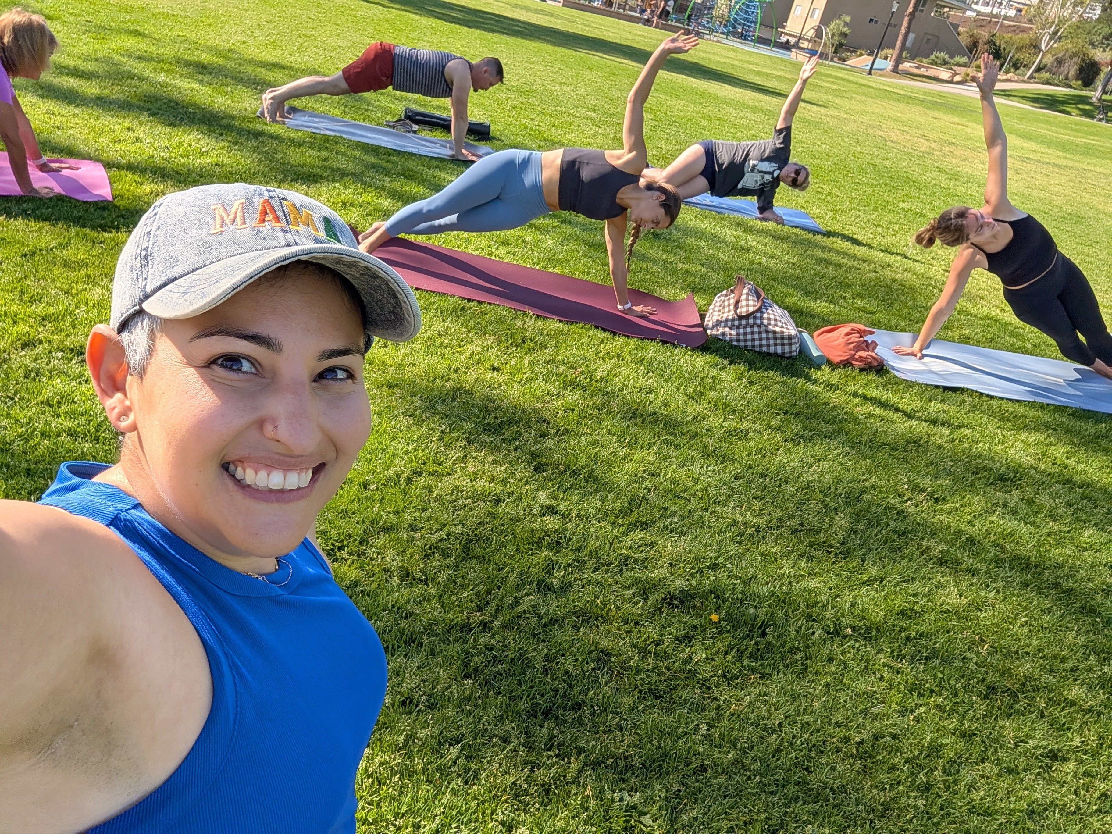 Young woman taking a selfie outdoors with people doing yoga in the background on grass.