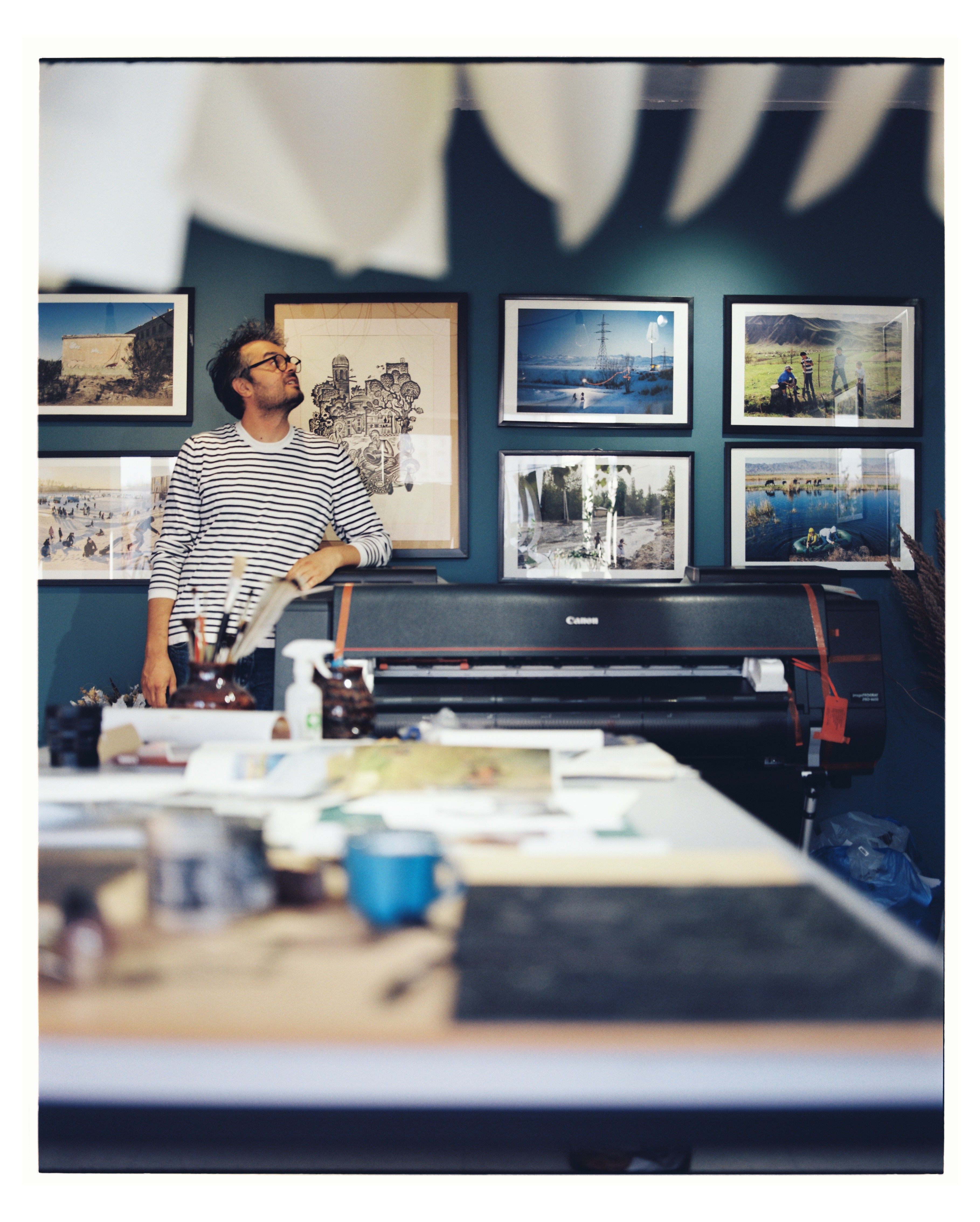 Person with glasses and striped shirt in art gallery, surrounded by framed photographs on dark blue wall, with a table in foreground.