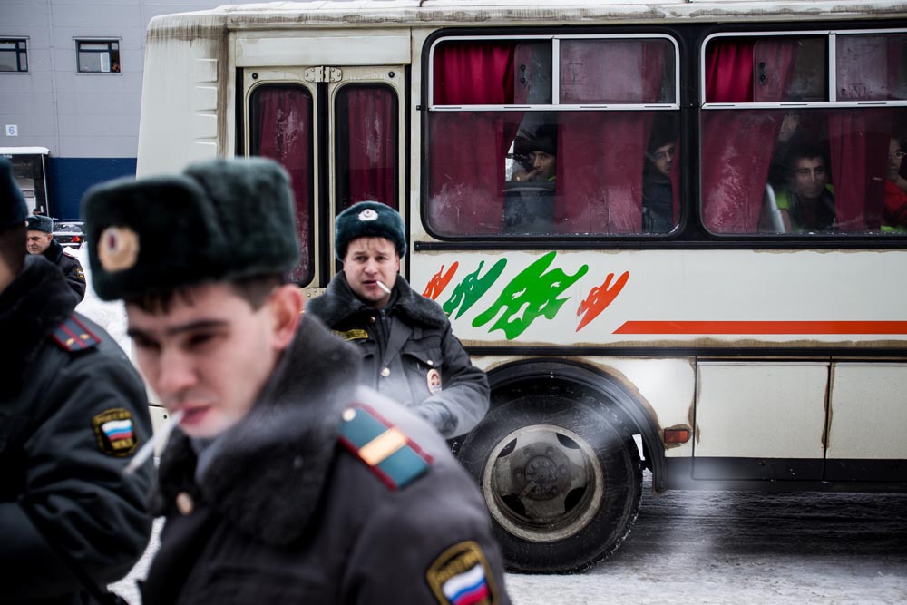 Two uniformed officers near a bus with red curtains, one in the foreground and one in the background, in an outdoor setting.