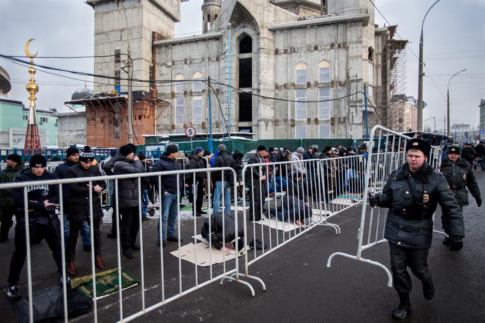 Crowd of people behind metal barricades near a large, partially constructed building under cloudy sky, with police officers present.