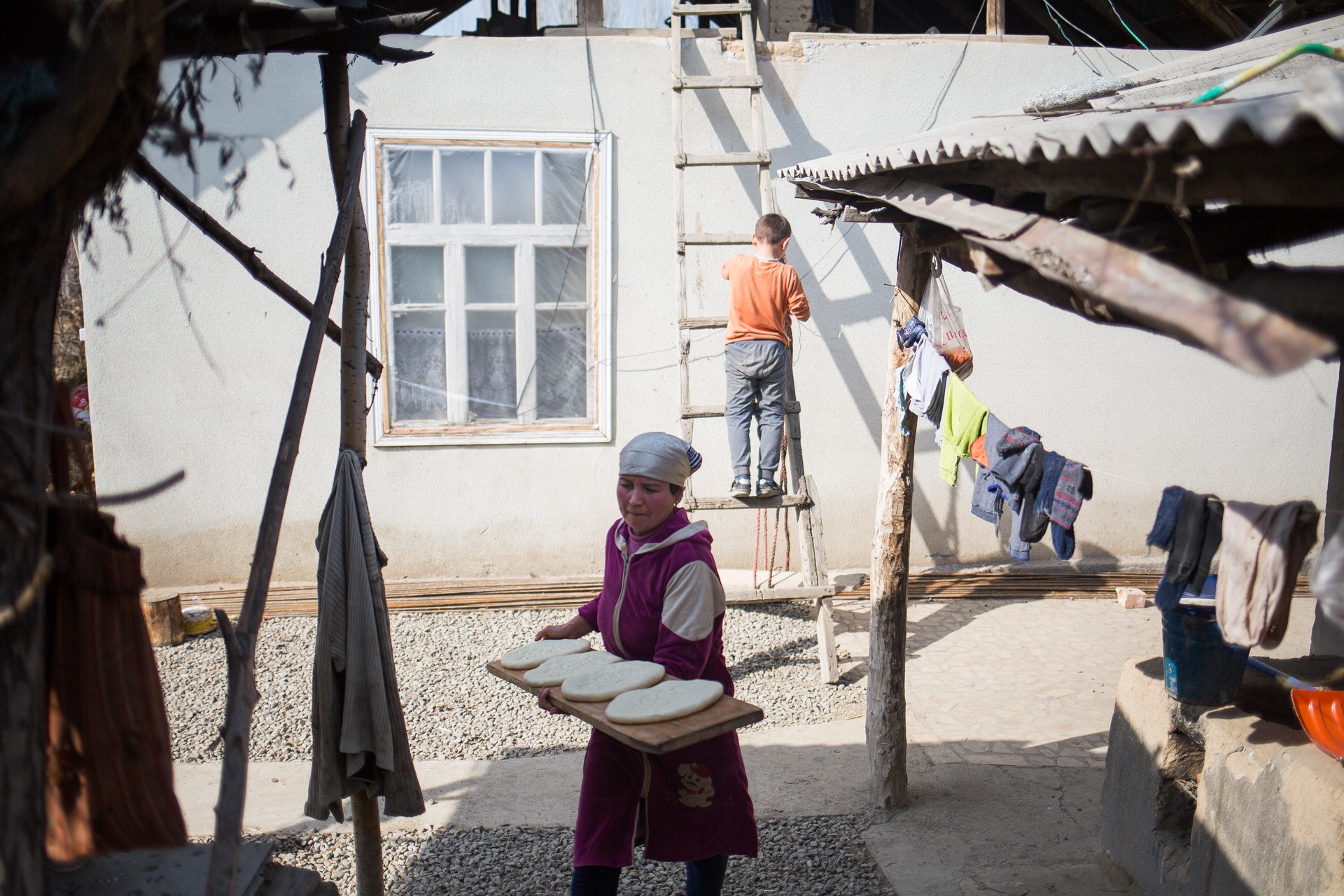 Child wearing a purple outfit and head covering holding trays outdoors near a building with a window.