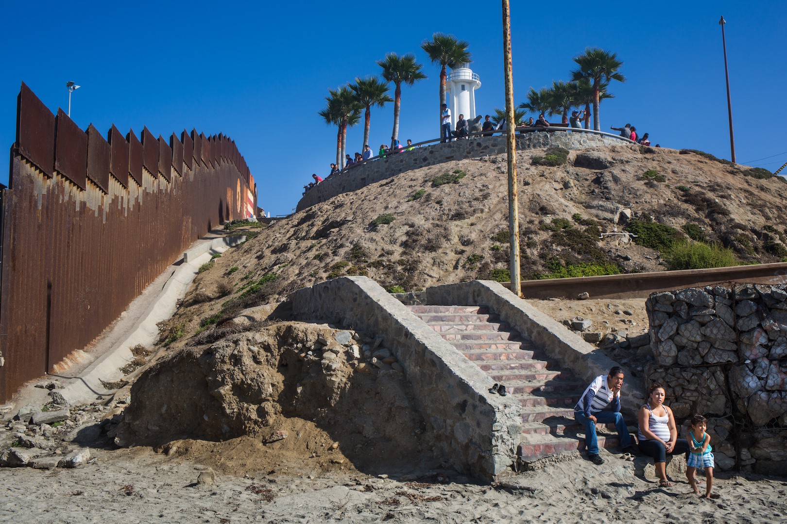 People sitting on stairs near a rocky hill with a lighthouse and palm trees on top, blue sky background.