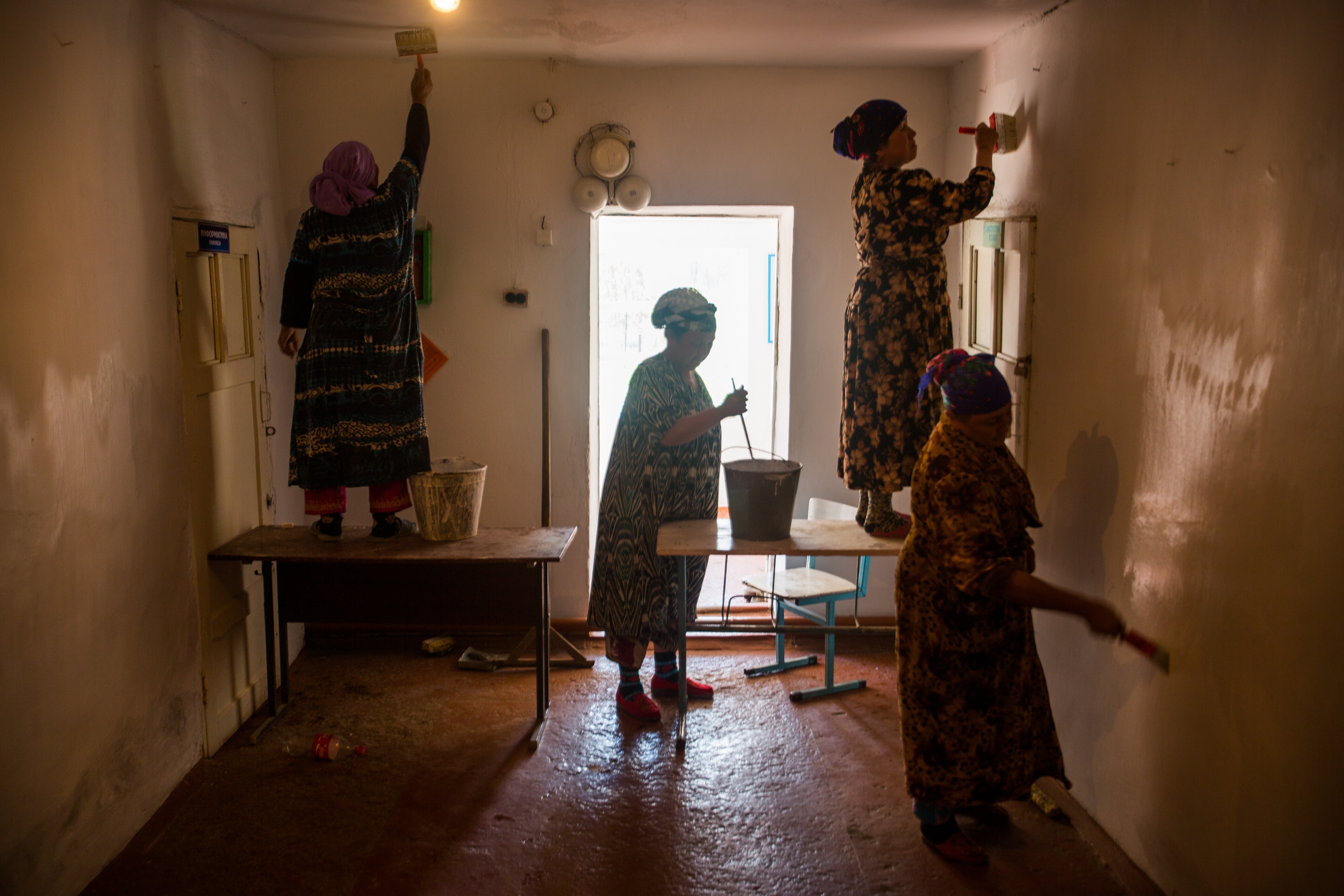 Four women in patterned dresses clean and dust a dimly lit room with a window, table, and bucket, backlit by sunlight.