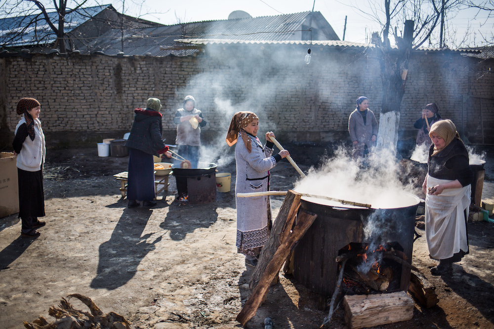 People gather outdoors around a large cooking pot over an open fire, some stirring or tending the food, with a brick wall in background.