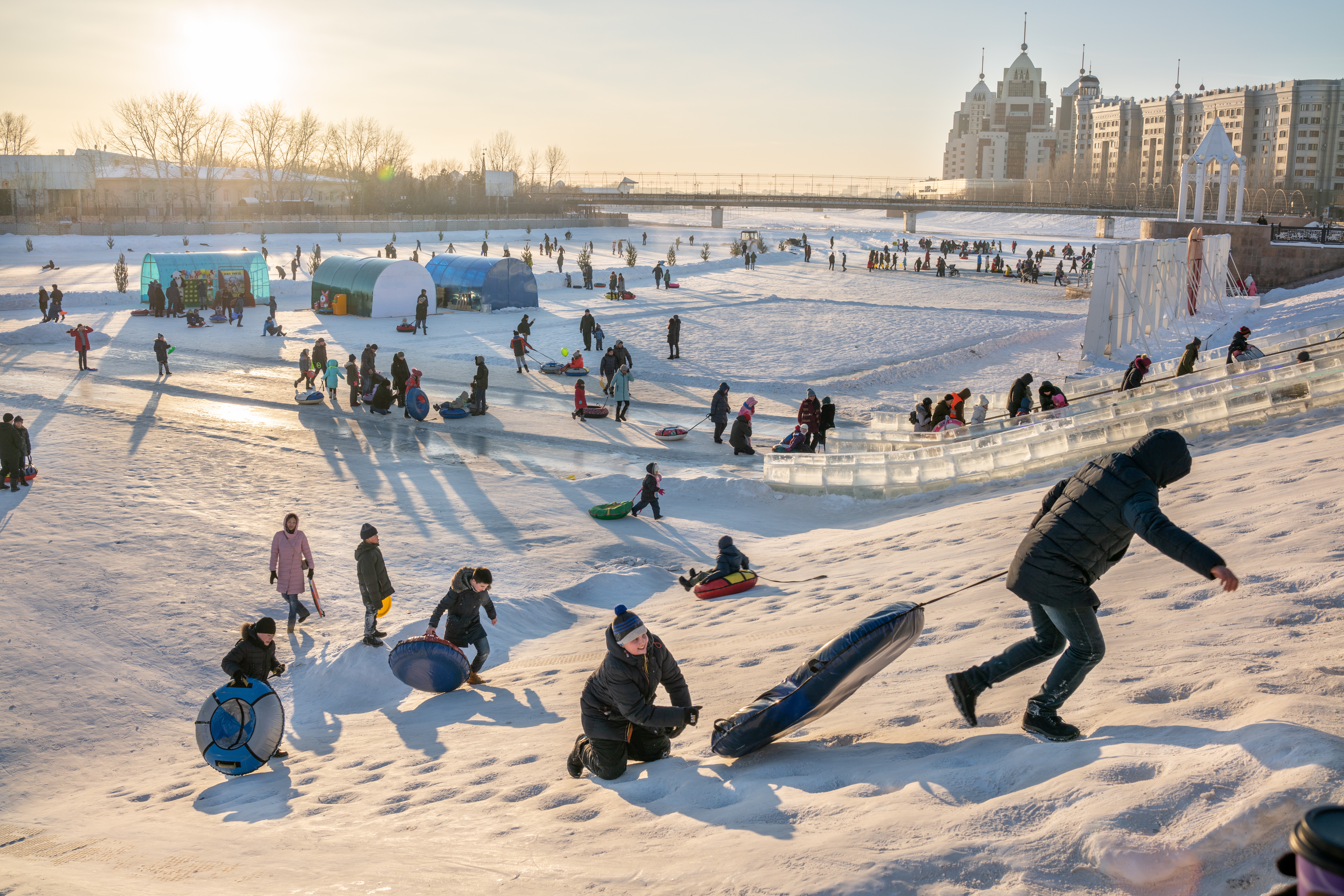 People sledding and playing on snow-covered hill with city buildings in background, winter scene, sunny day.