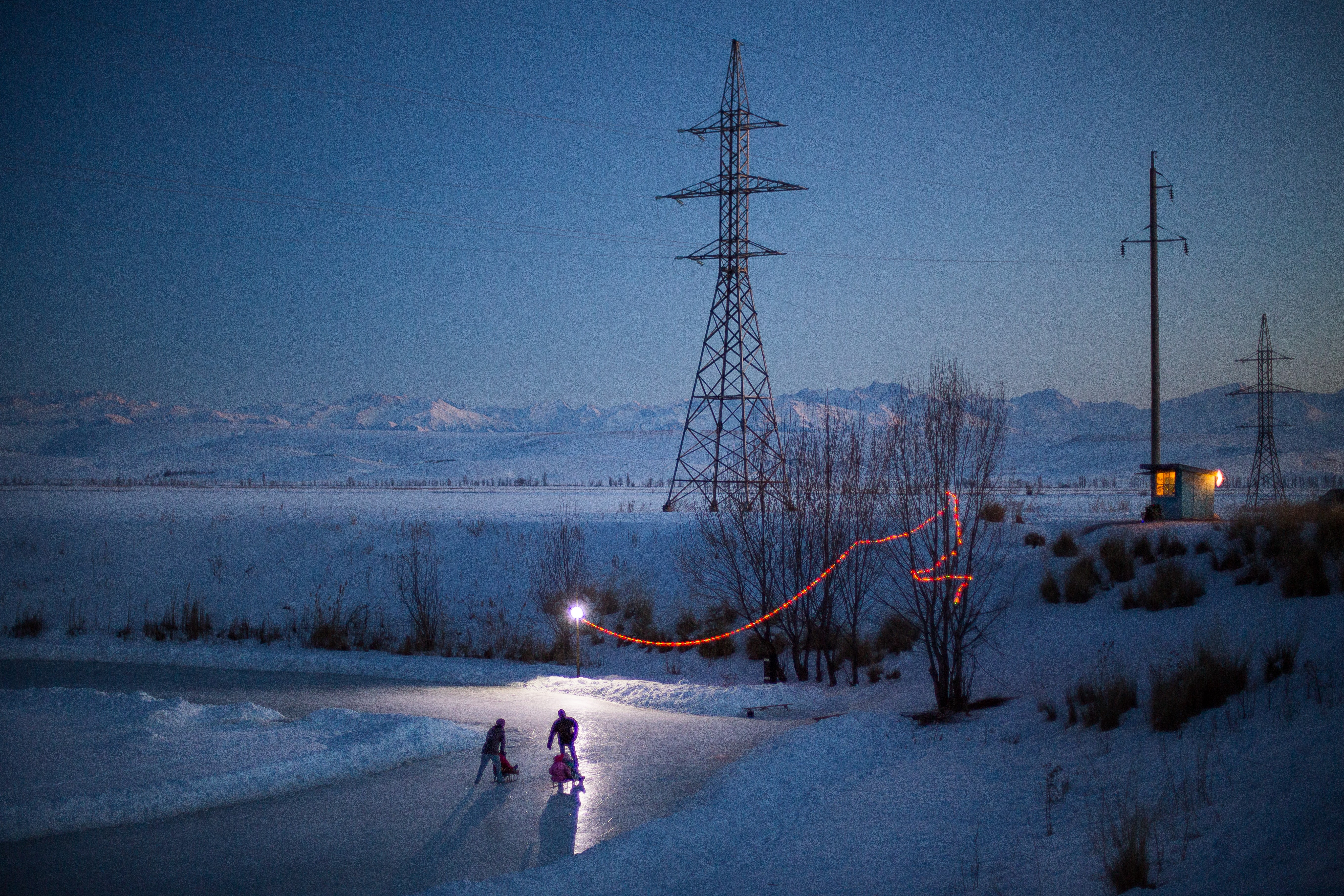 Snow-covered landscape with power lines, a small building, and two children walking in the snow at dusk.