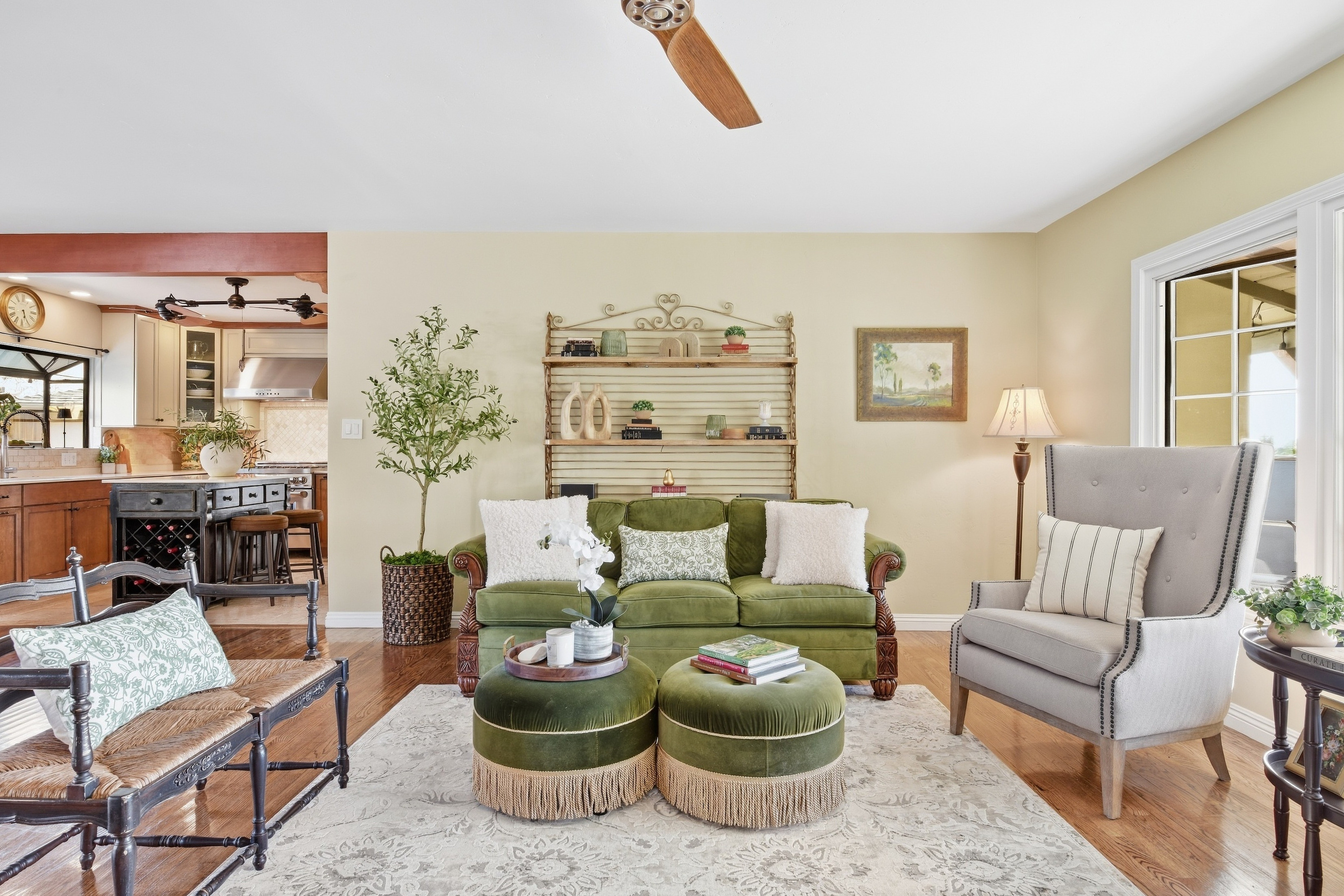 Living room with green sofa, armchair, ottomans, and a ceiling fan, with a view into the kitchen area, and a large window.