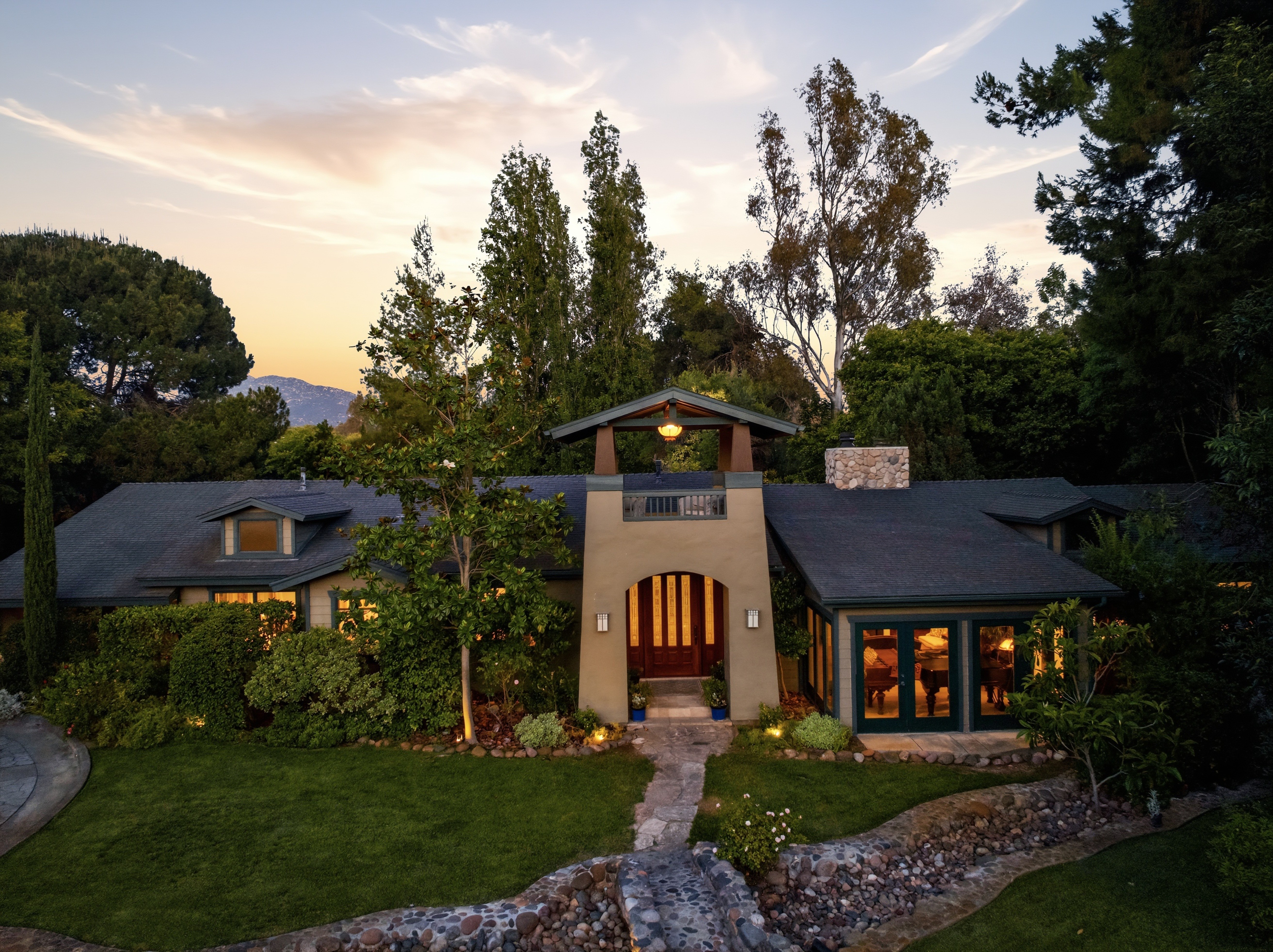 House with a central entrance, surrounded by trees and greenery, under a partly cloudy sky at sunset.