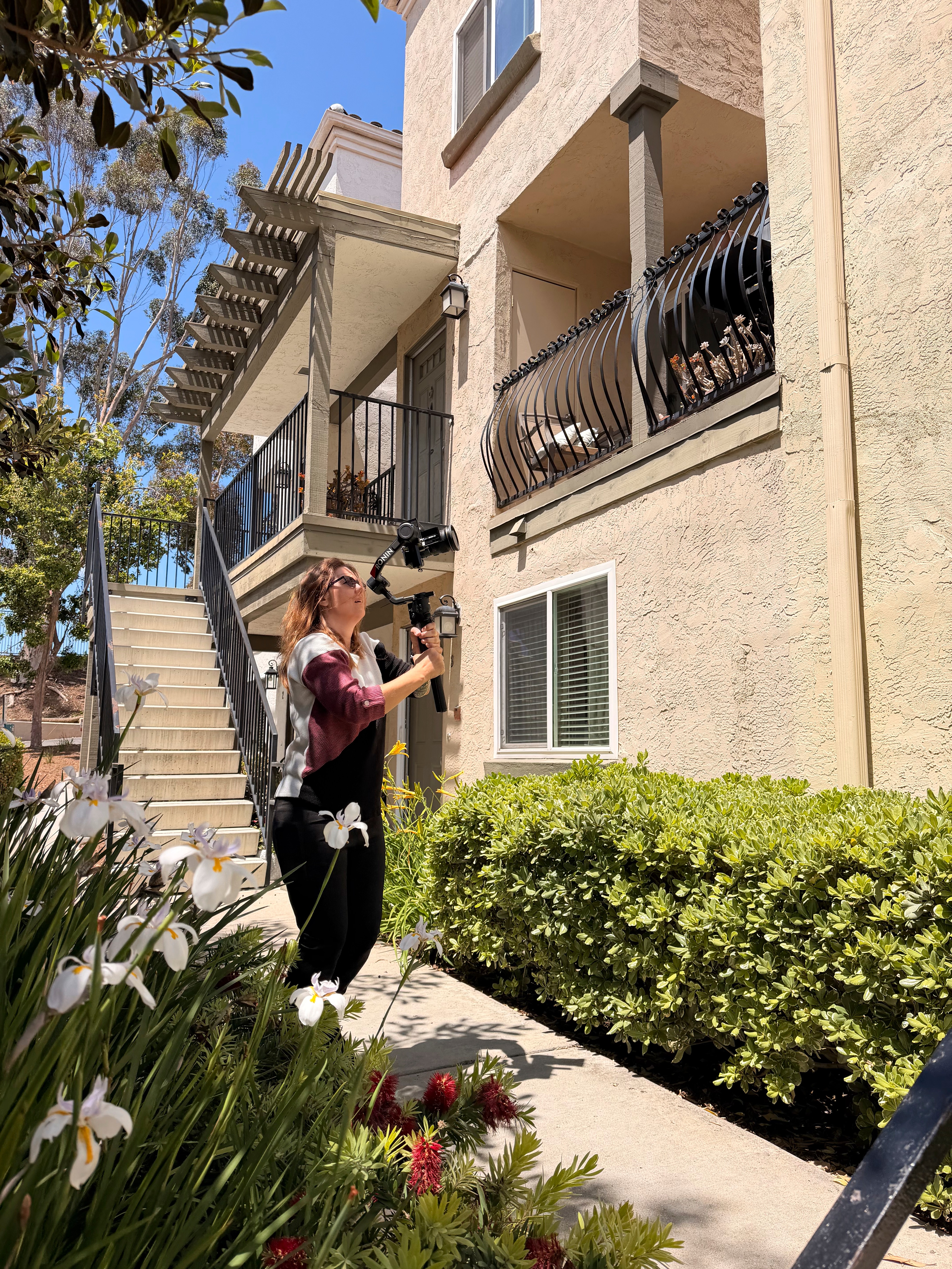 Woman standing outside near bushes, taking a photo with a smartphone, building with balconies in background.