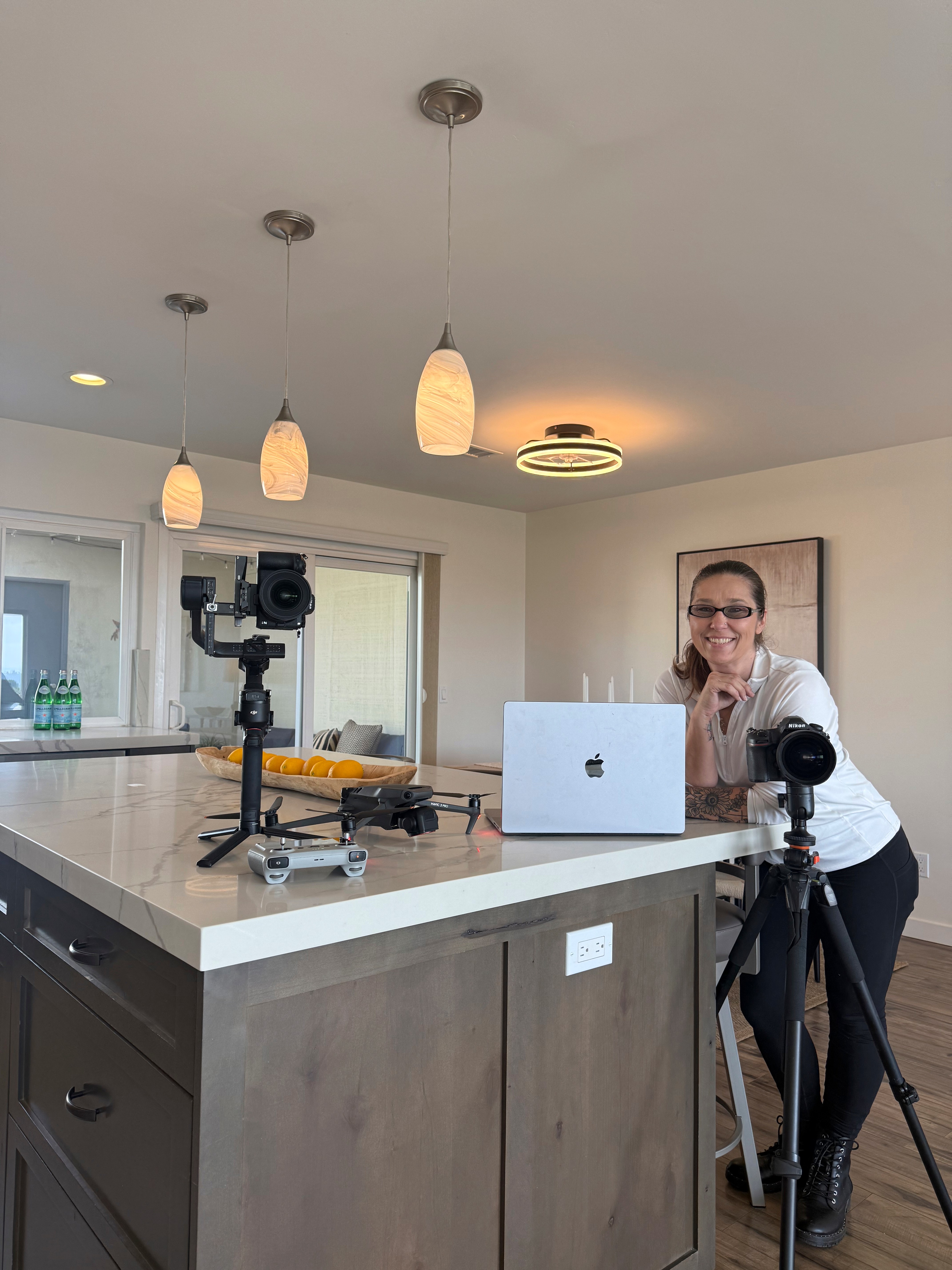 Woman smiling at a kitchen island with a laptop, camera, and tripod, in a well-lit room with hanging lights.