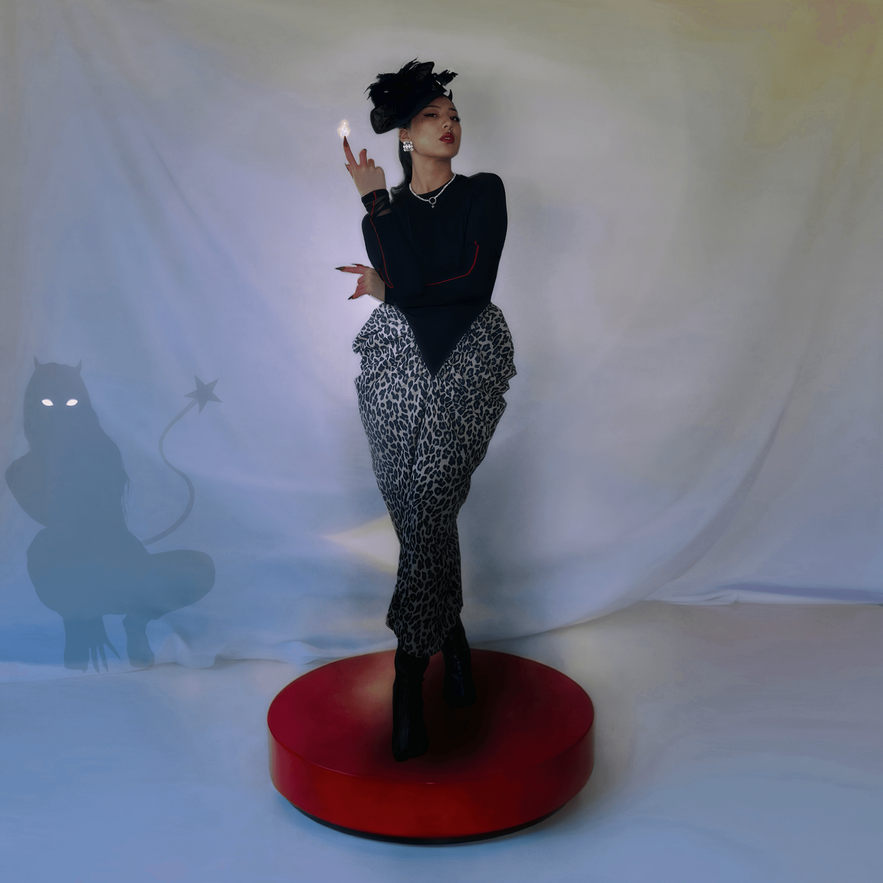 Woman standing on a red platform, wearing leopard print pants and a black top, with a dark background and a cat silhouette.