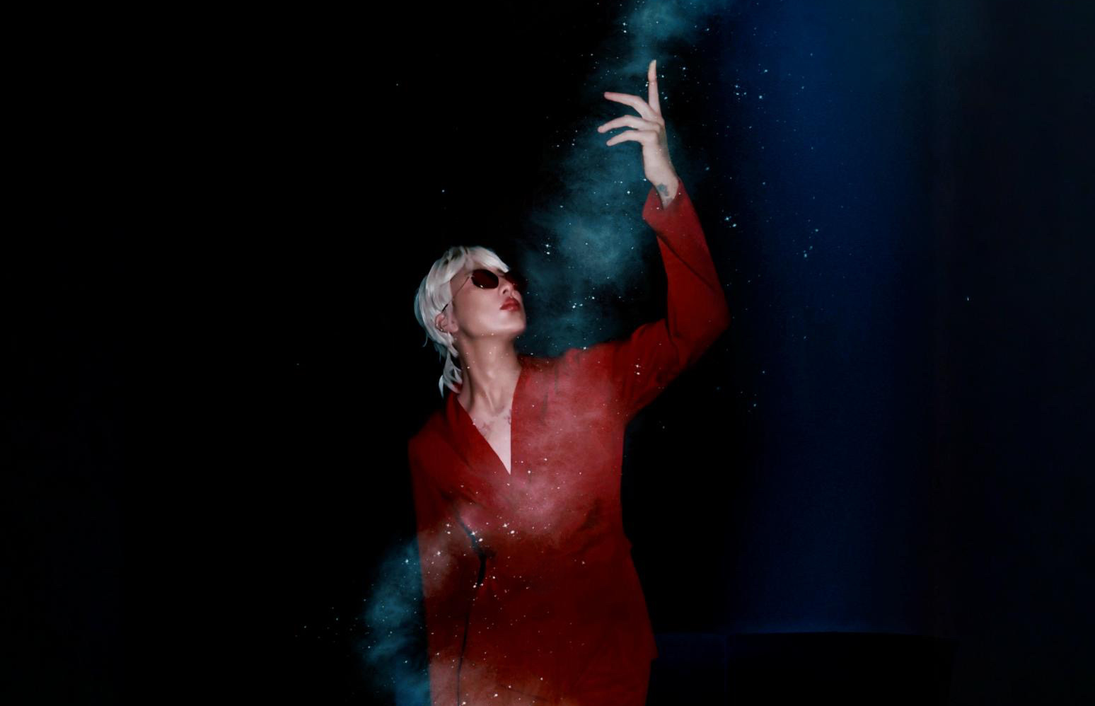 Person with short blonde hair and sunglasses reaches upward, with a blue powder cloud around their hand, wearing a red outfit.