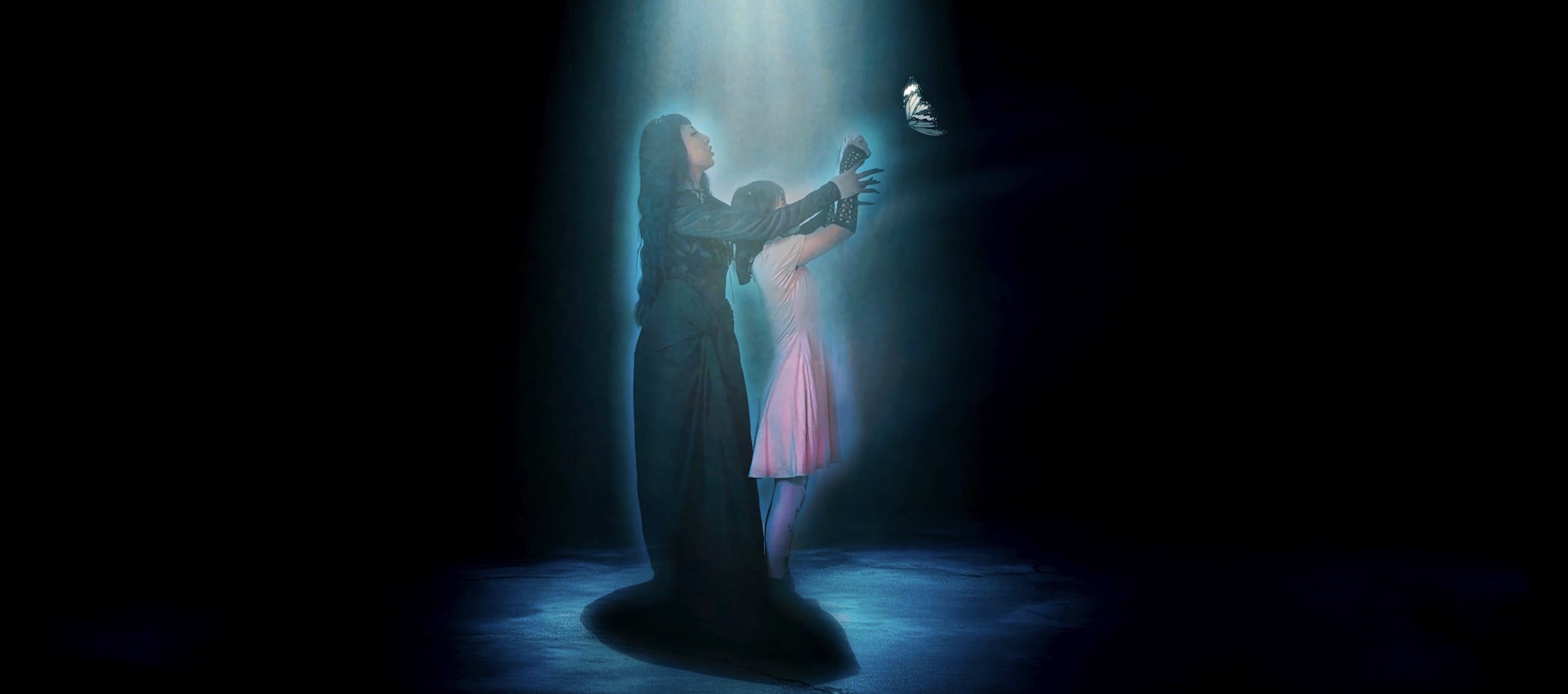 A person kneeling and holding a child in a dark setting with blue lighting, both facing each other.