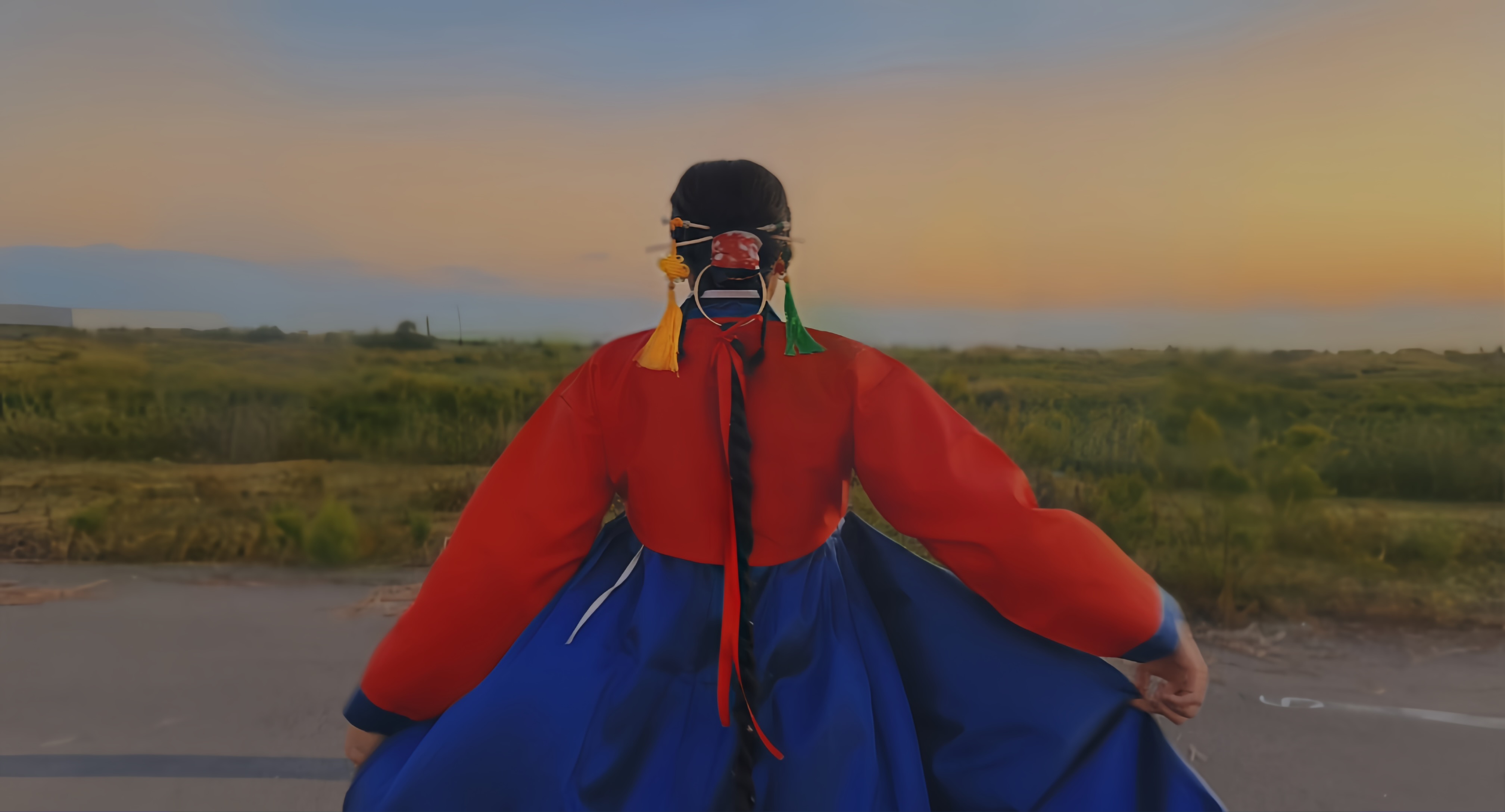 Person in traditional clothing standing outdoors with a landscape and sunset in the background.