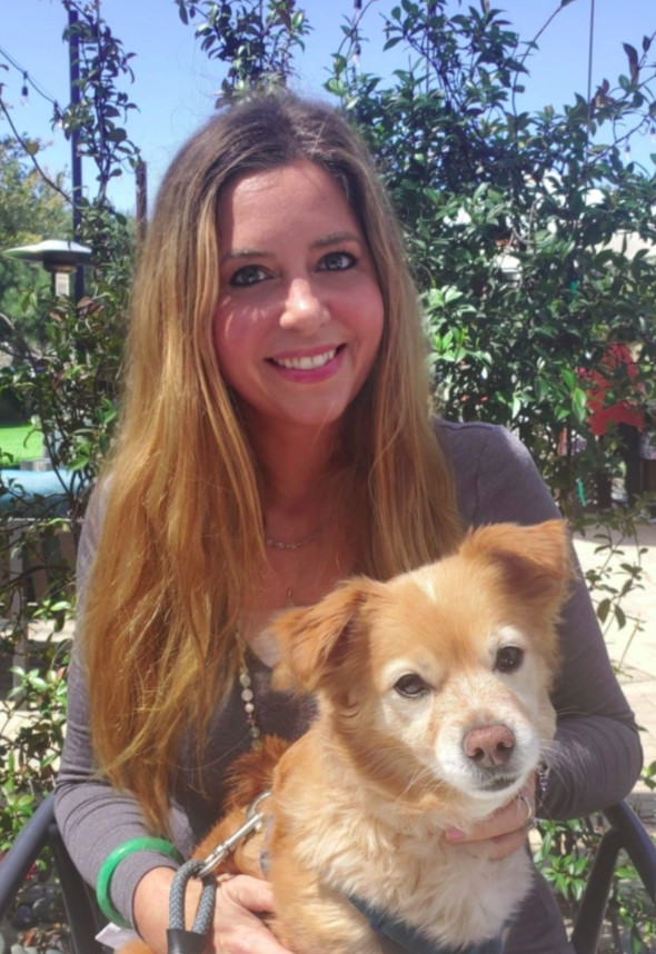 Woman with long hair smiling outdoors holding a small tan and white dog, trees and sky in background.
