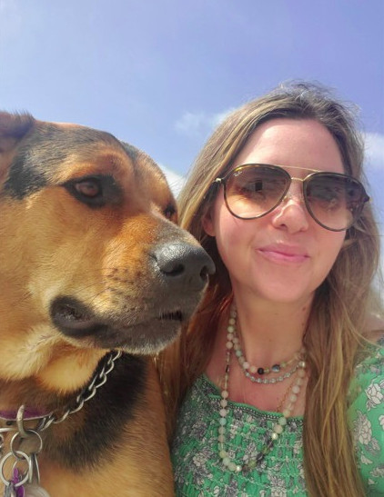 Woman with long hair wearing sunglasses and a green patterned top, smiling next to a large dog outdoors under a blue sky.