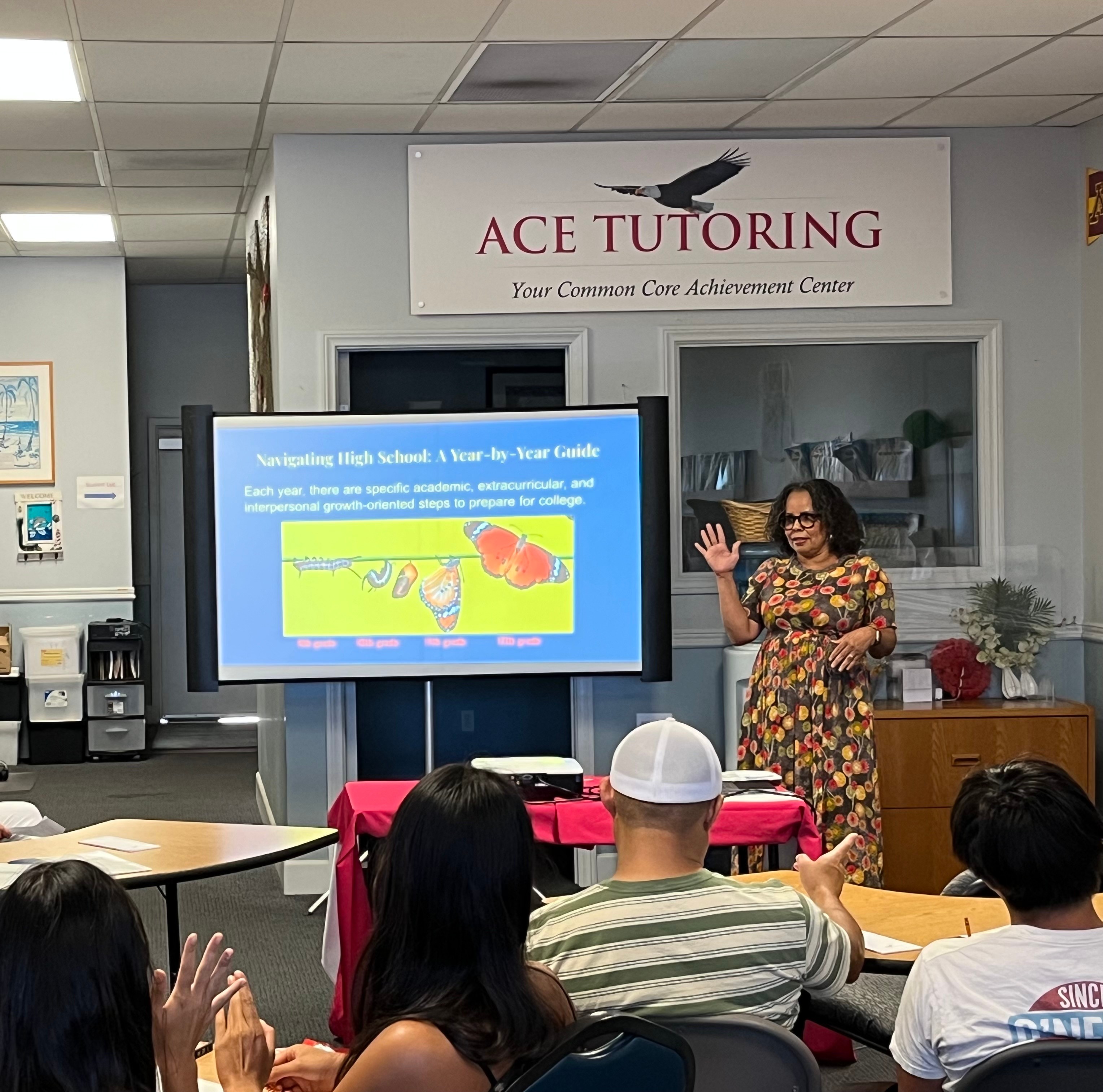A woman stands in front of a classroom, gesturing with her right hand, with students seated and clapping, in a tutoring center.