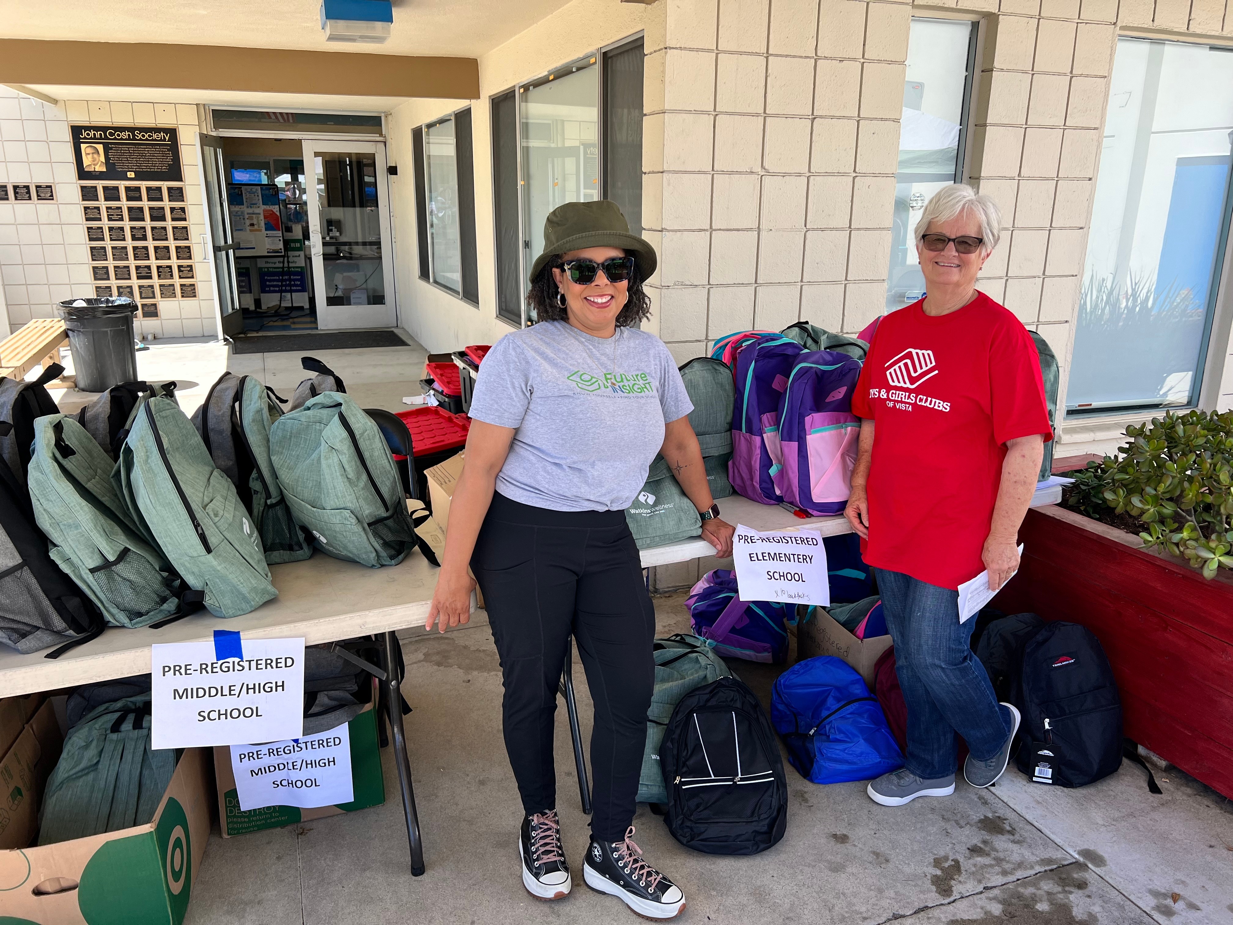 Two women standing outdoors with backpacks and supplies, one wearing a hat and sunglasses, the other in a red shirt, in front of a building.