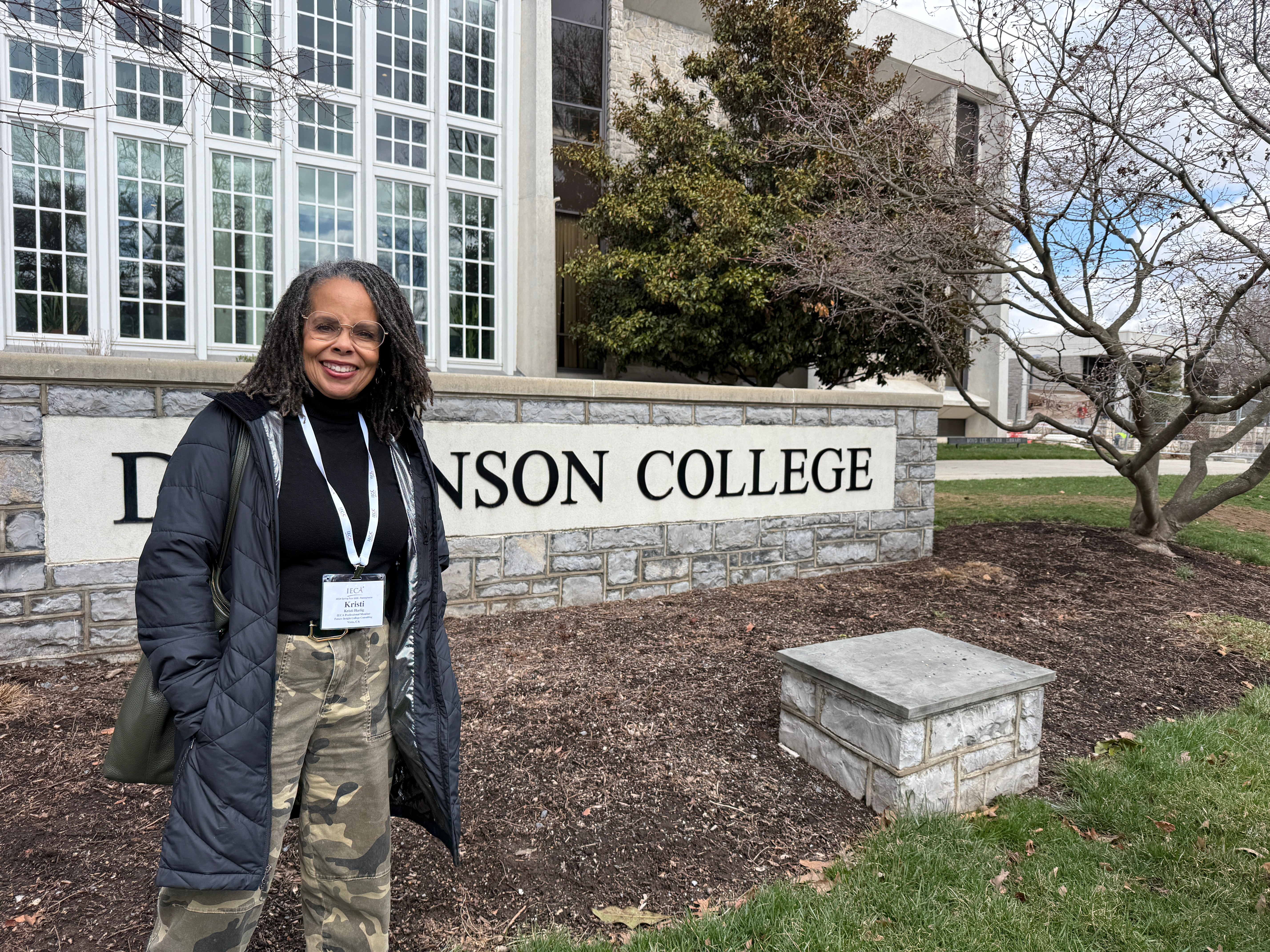 Smiling woman standing outdoors near a college sign, wearing a black jacket and camouflage pants, with trees and a building in the background.