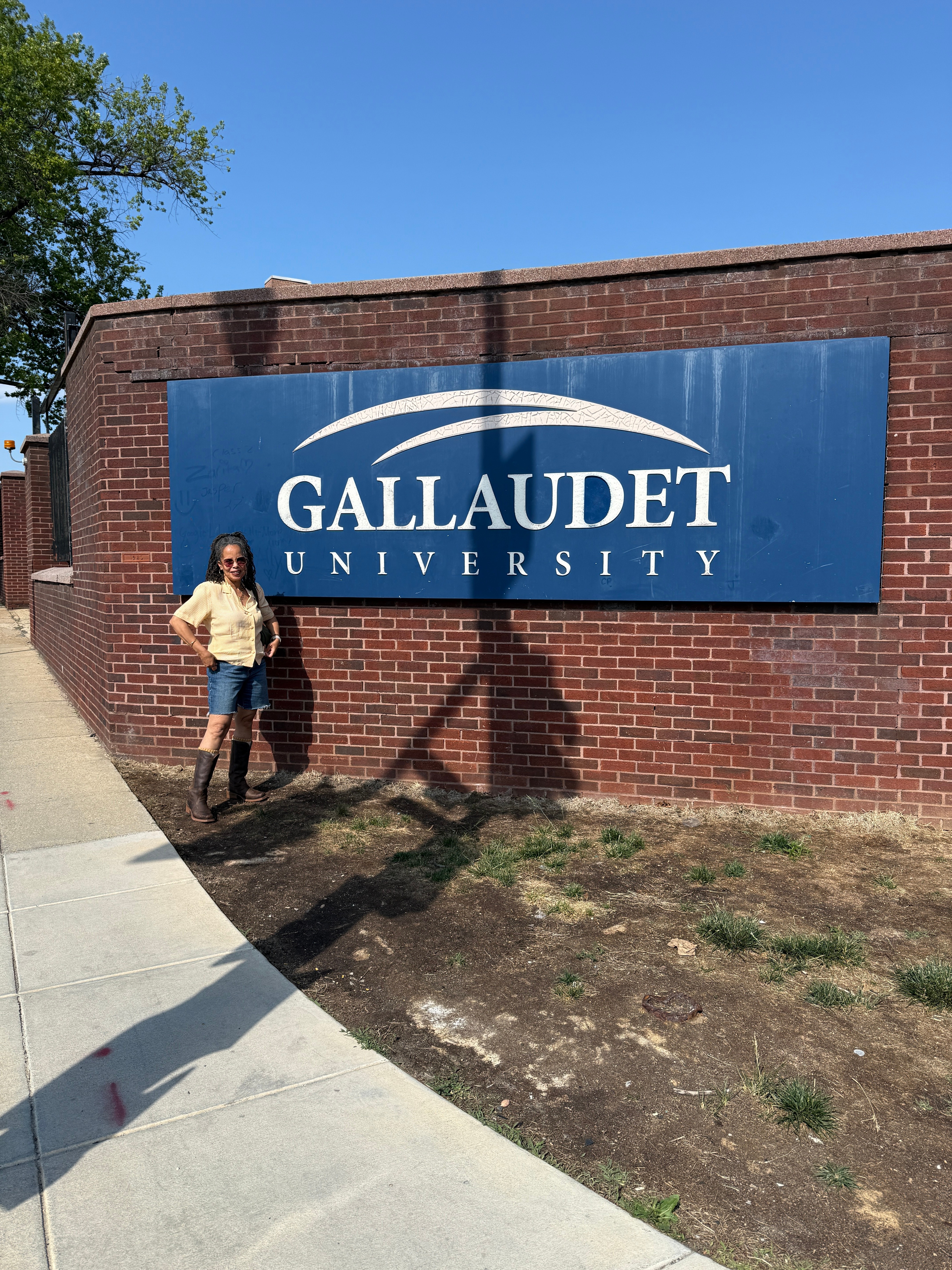 Person standing next to a Gallaudet University sign on a brick wall, with a clear blue sky above.