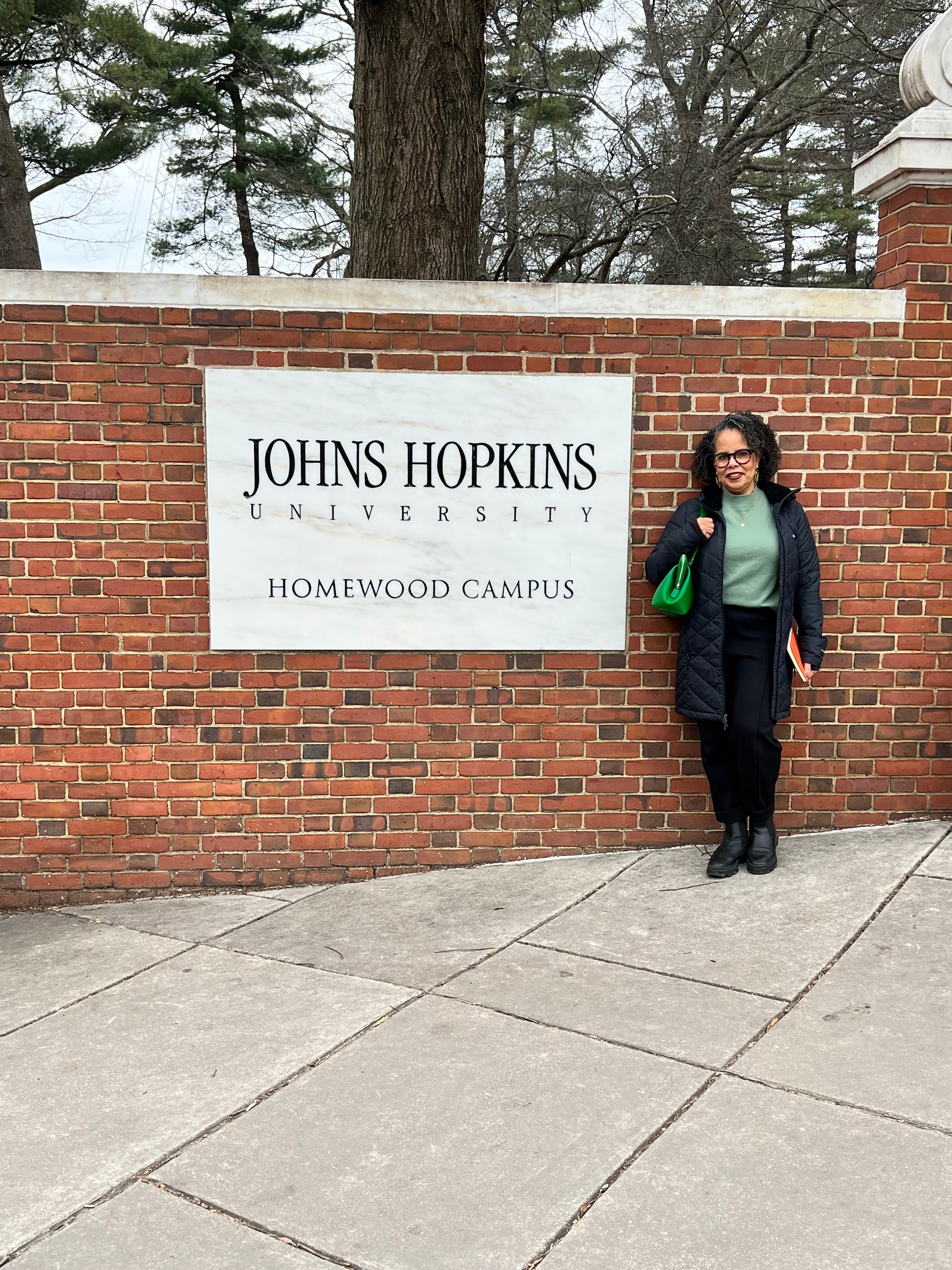 Woman standing next to a brick wall with a sign for Johns Hopkins University Homewood Campus, outdoors, trees in background.