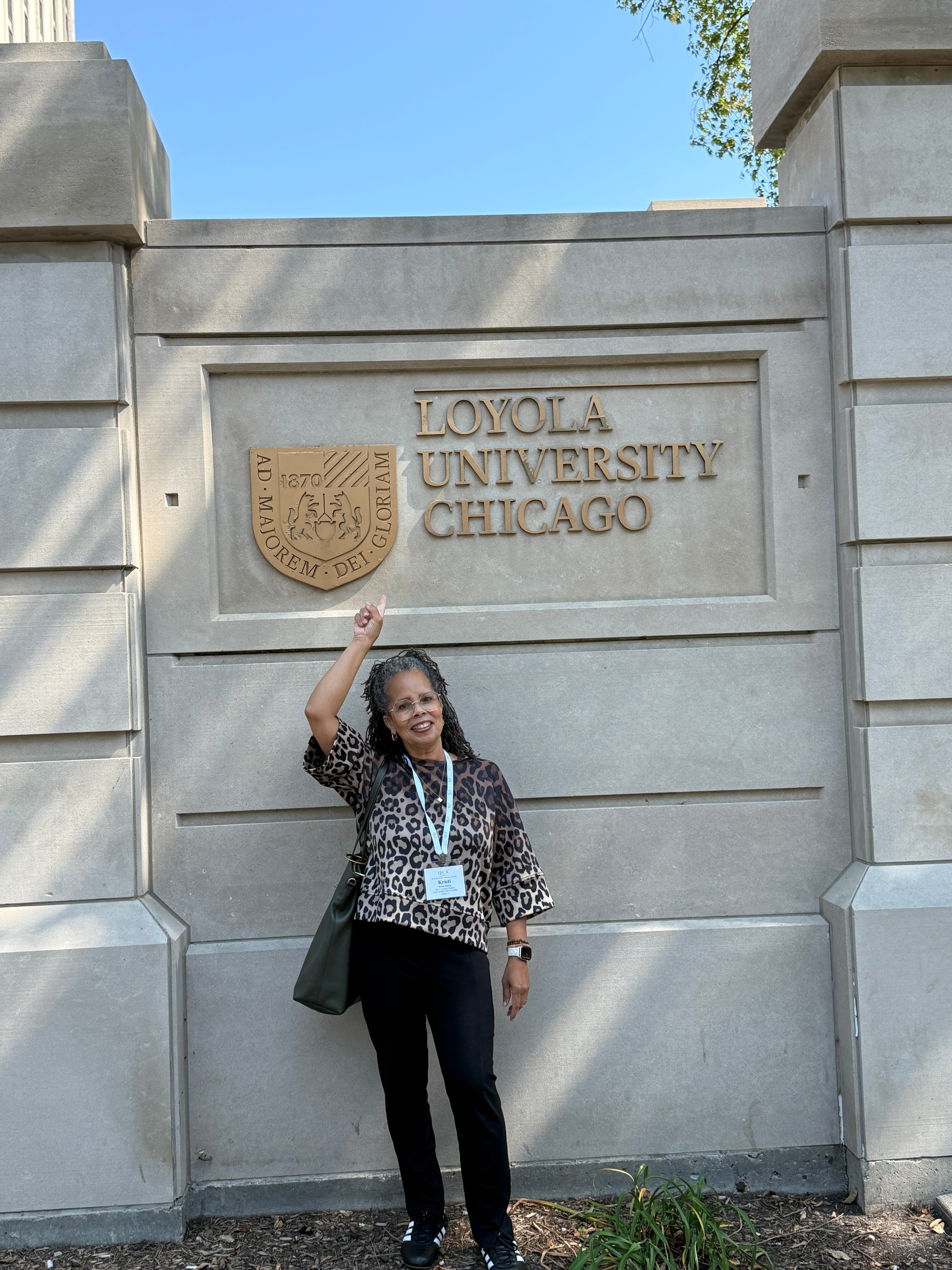 Woman standing in front of Loyola University Chicago sign, smiling and waving, wearing a leopard print top and black pants.