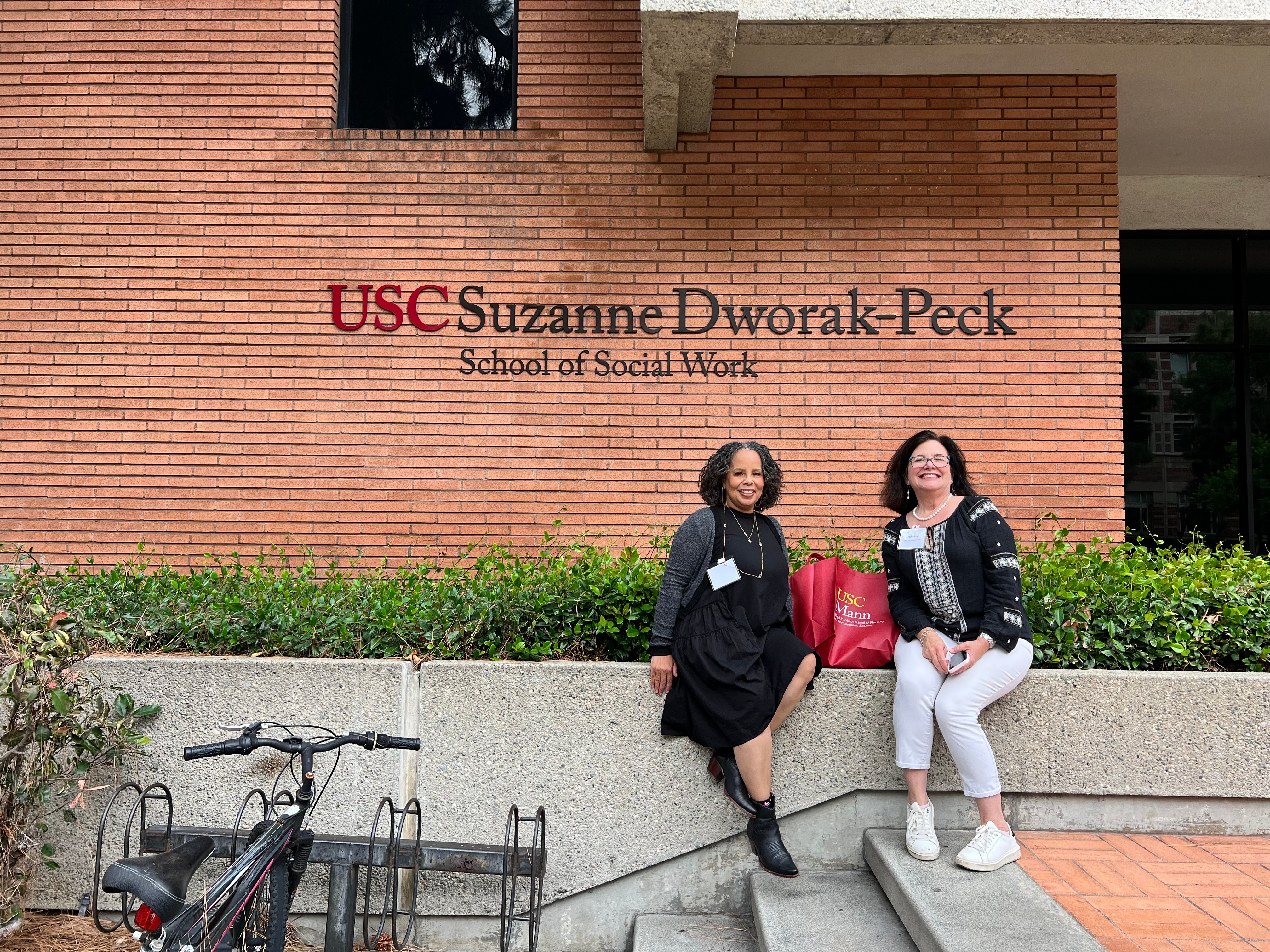 Two women sit on a concrete ledge in front of a brick building with a sign reading 'USC Suzanne Dworak-Peck School of Social Work.'