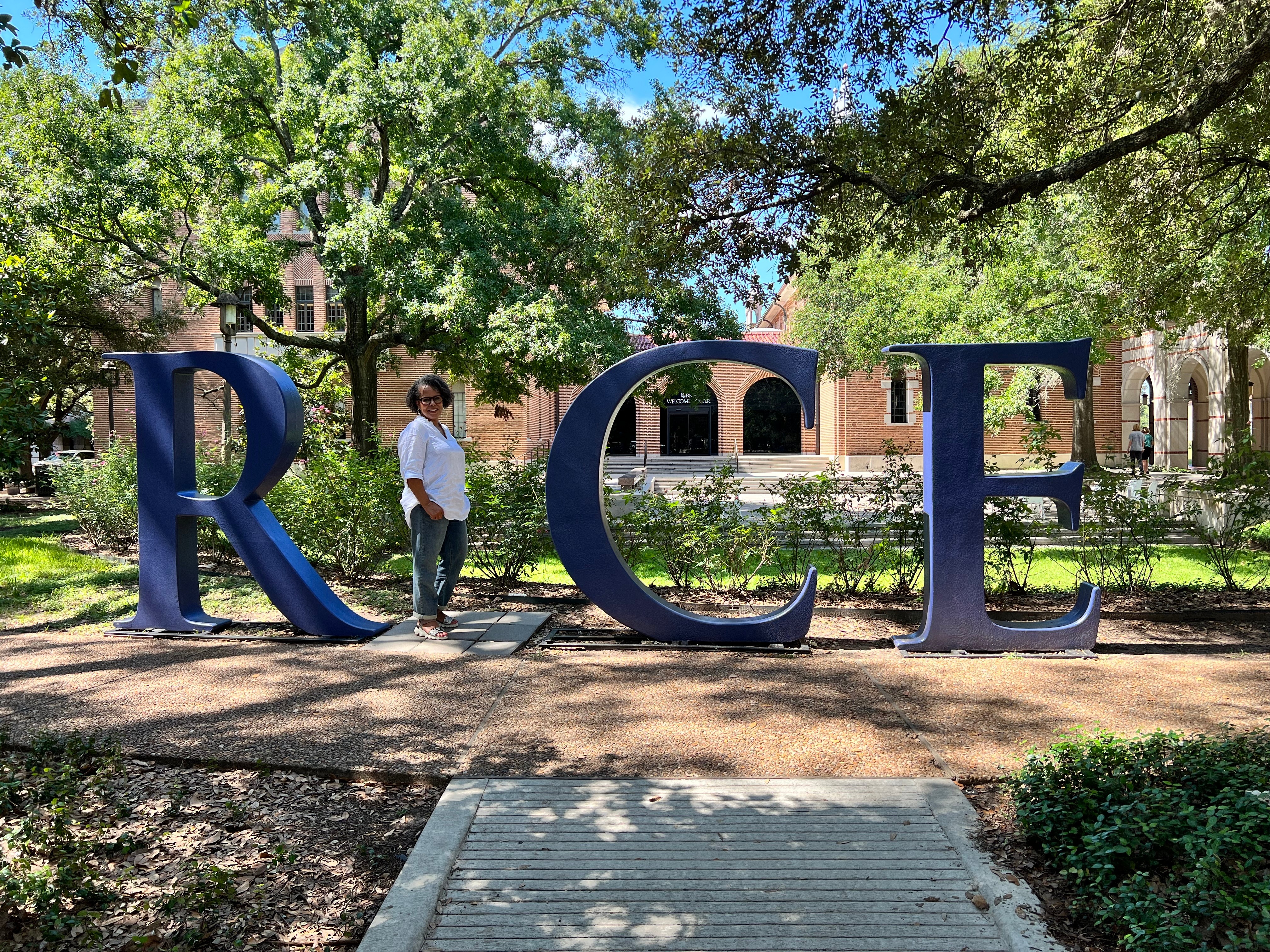 Person standing next to large blue letters spelling 'RICE' outdoors with trees and buildings in background.