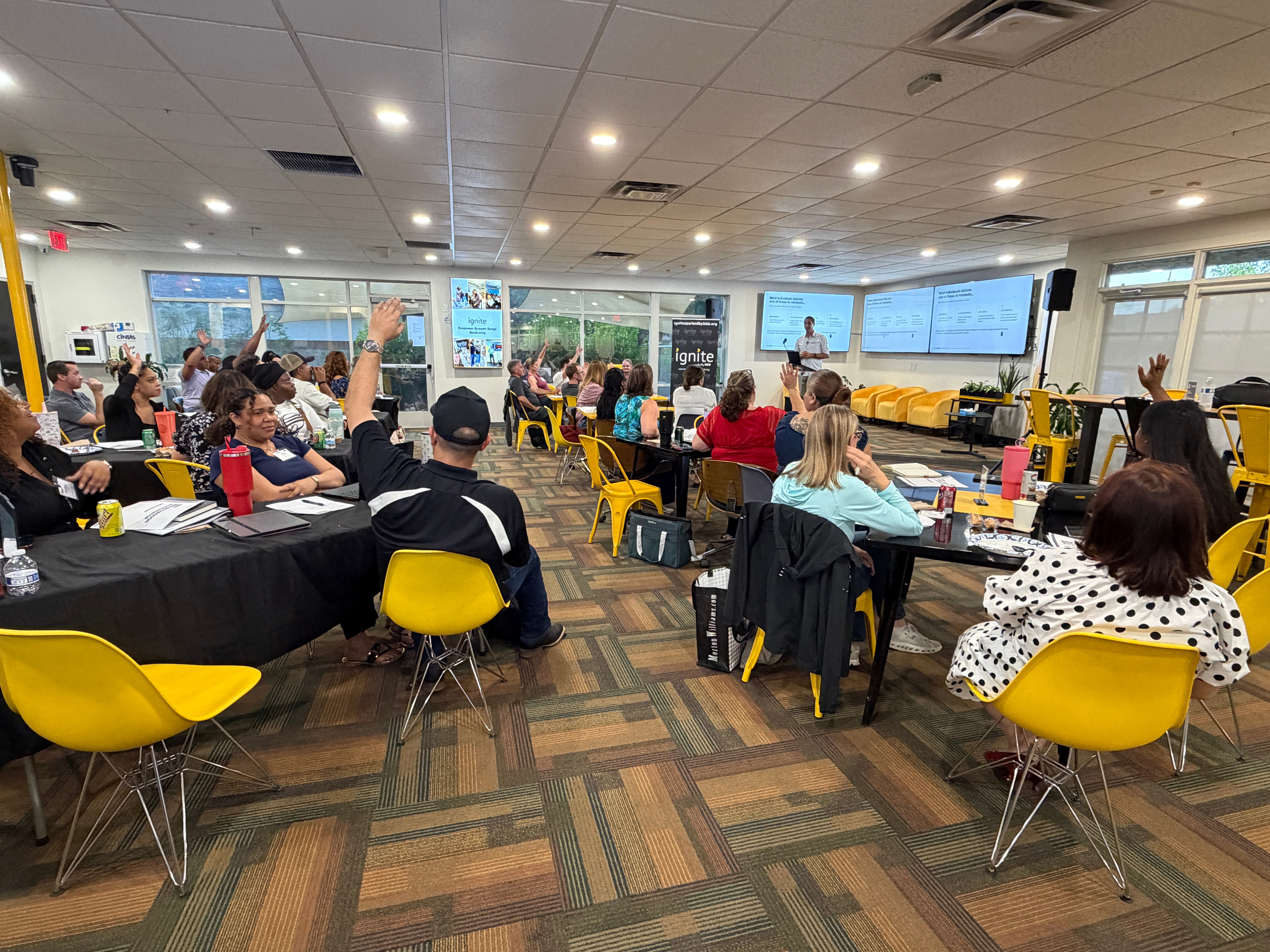 People seated at tables in a conference room with large screens and windows, some raising hands, yellow chairs, and a patterned floor.