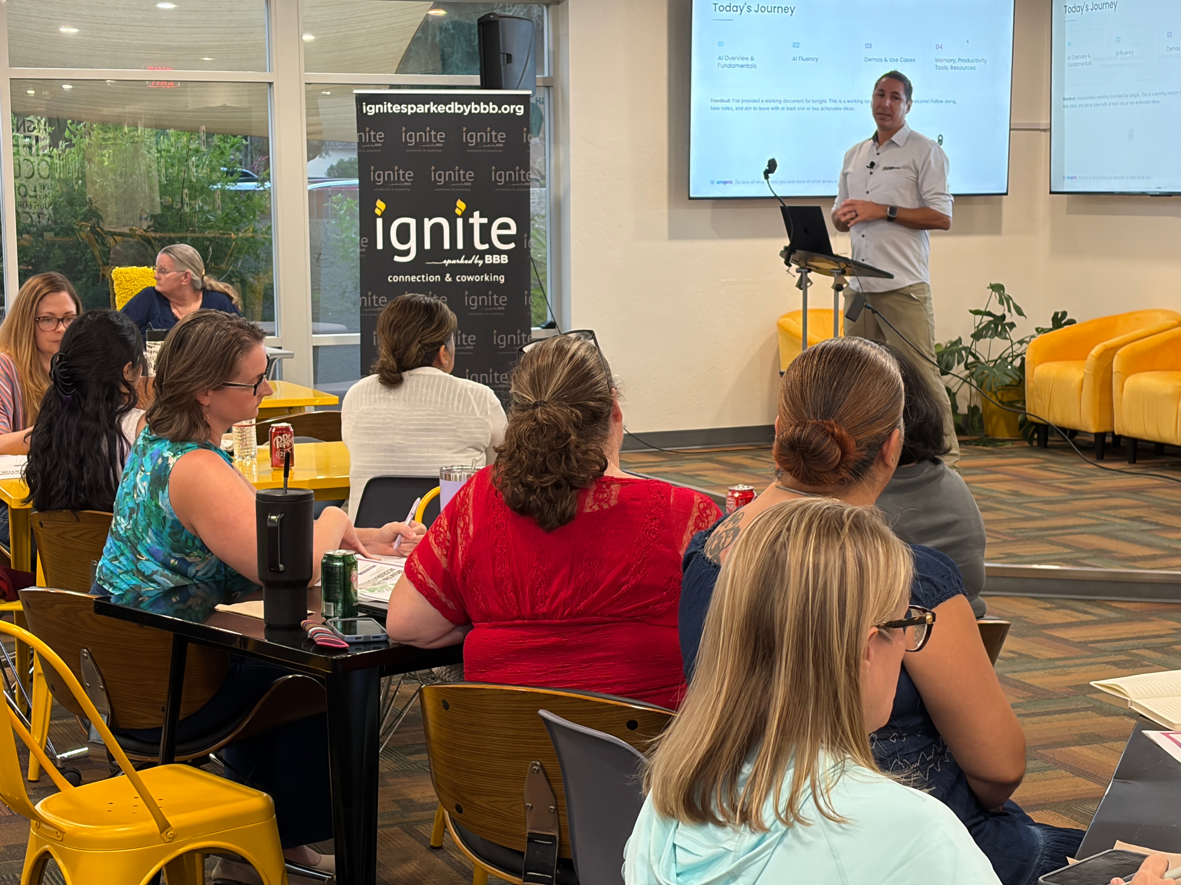 Person standing at podium giving presentation to seated audience in a modern room with large windows and yellow chairs.