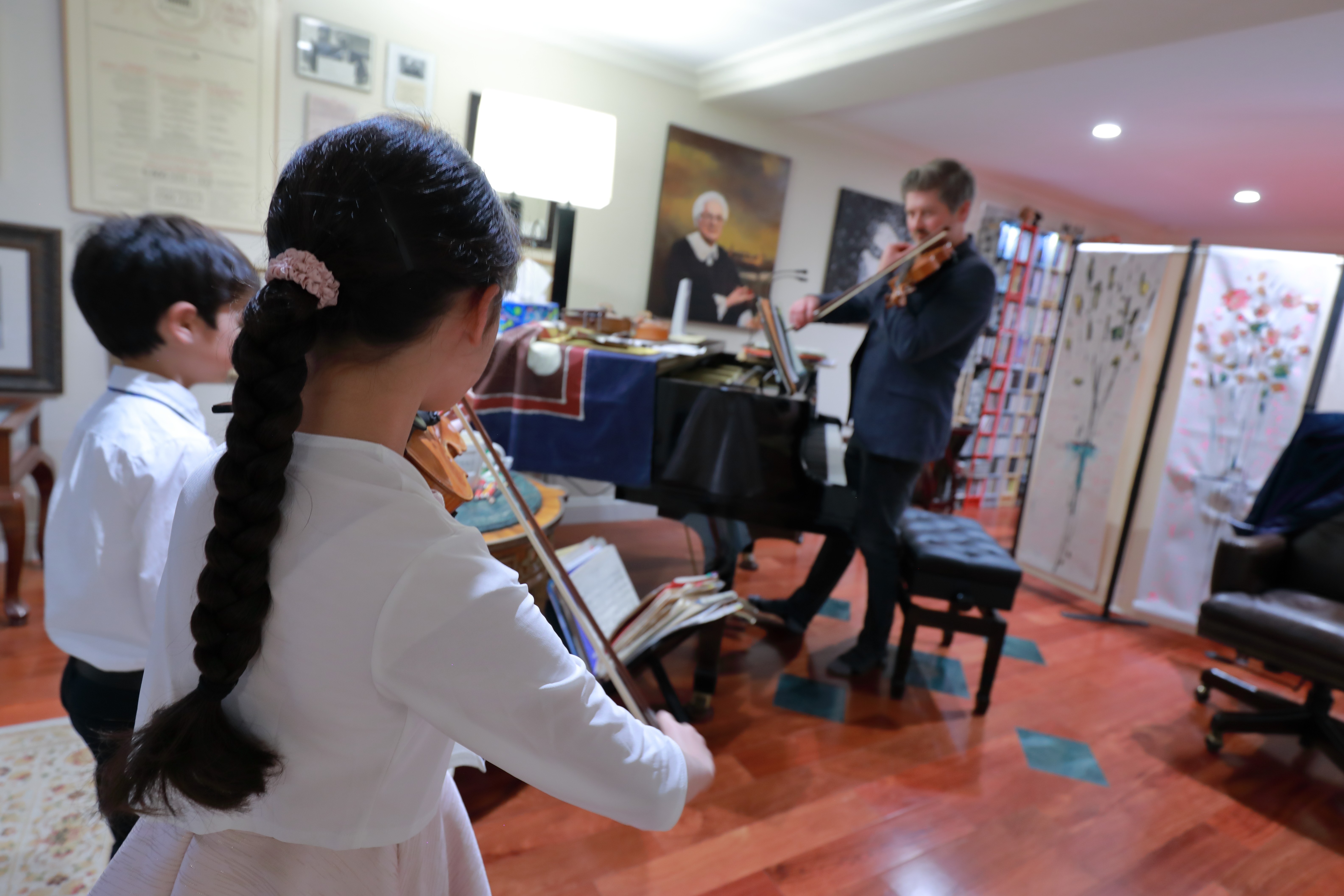Children playing violins while a person performs on a piano in a room with artwork and furniture.