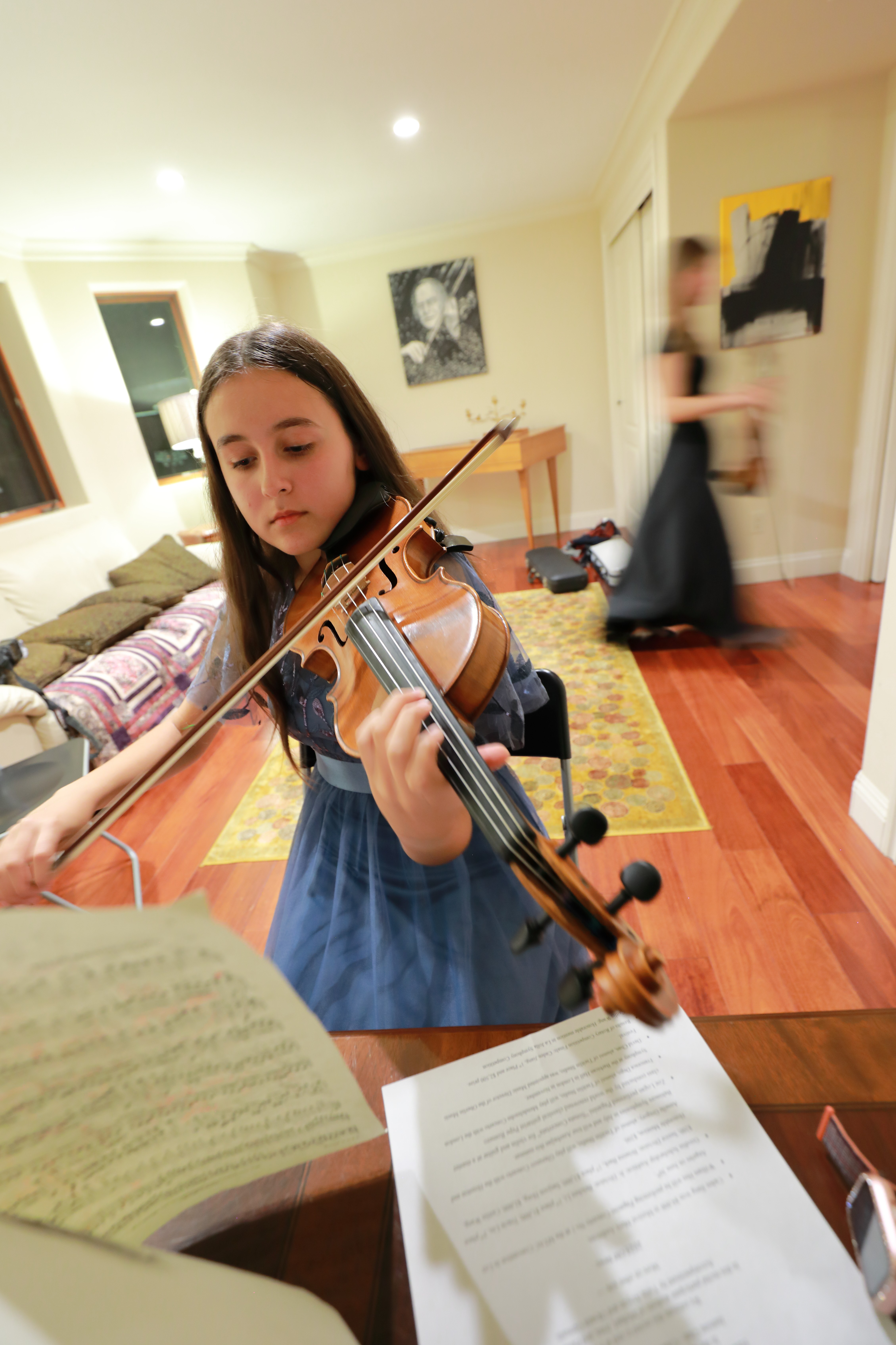 Young girl playing violin indoors, with sheet music in foreground, blurred person walking in background, warm lighting, wooden floor.