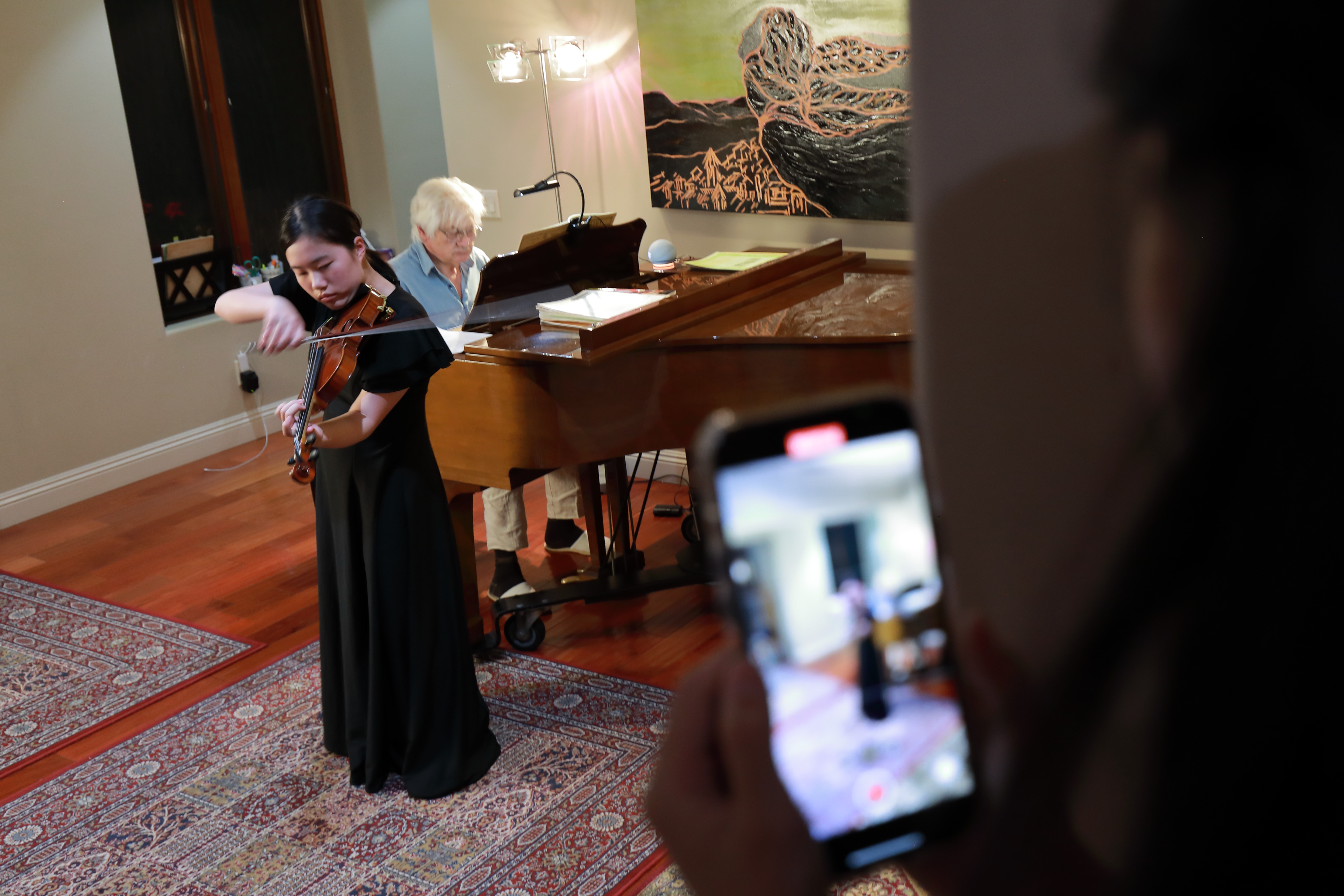 A young girl plays the violin while an older woman takes a photo with a smartphone in a room with a piano and artwork.