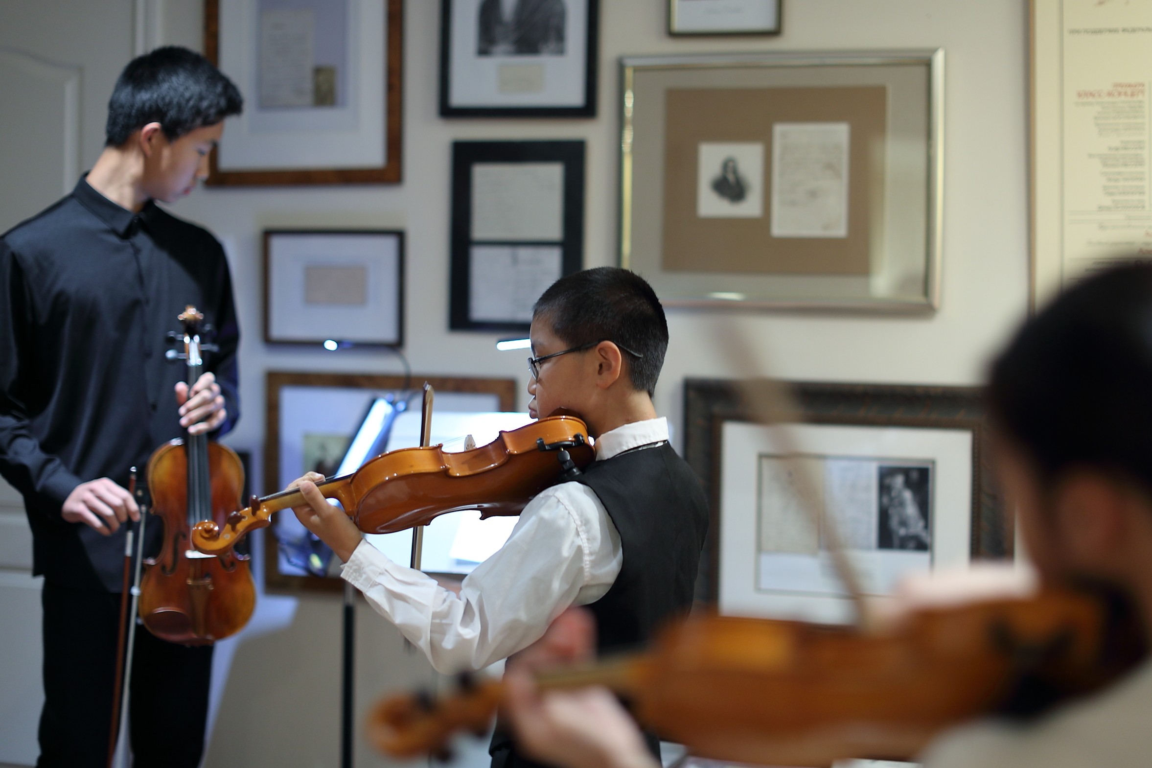 Two children playing violins in an art gallery with framed pictures on the wall behind them.