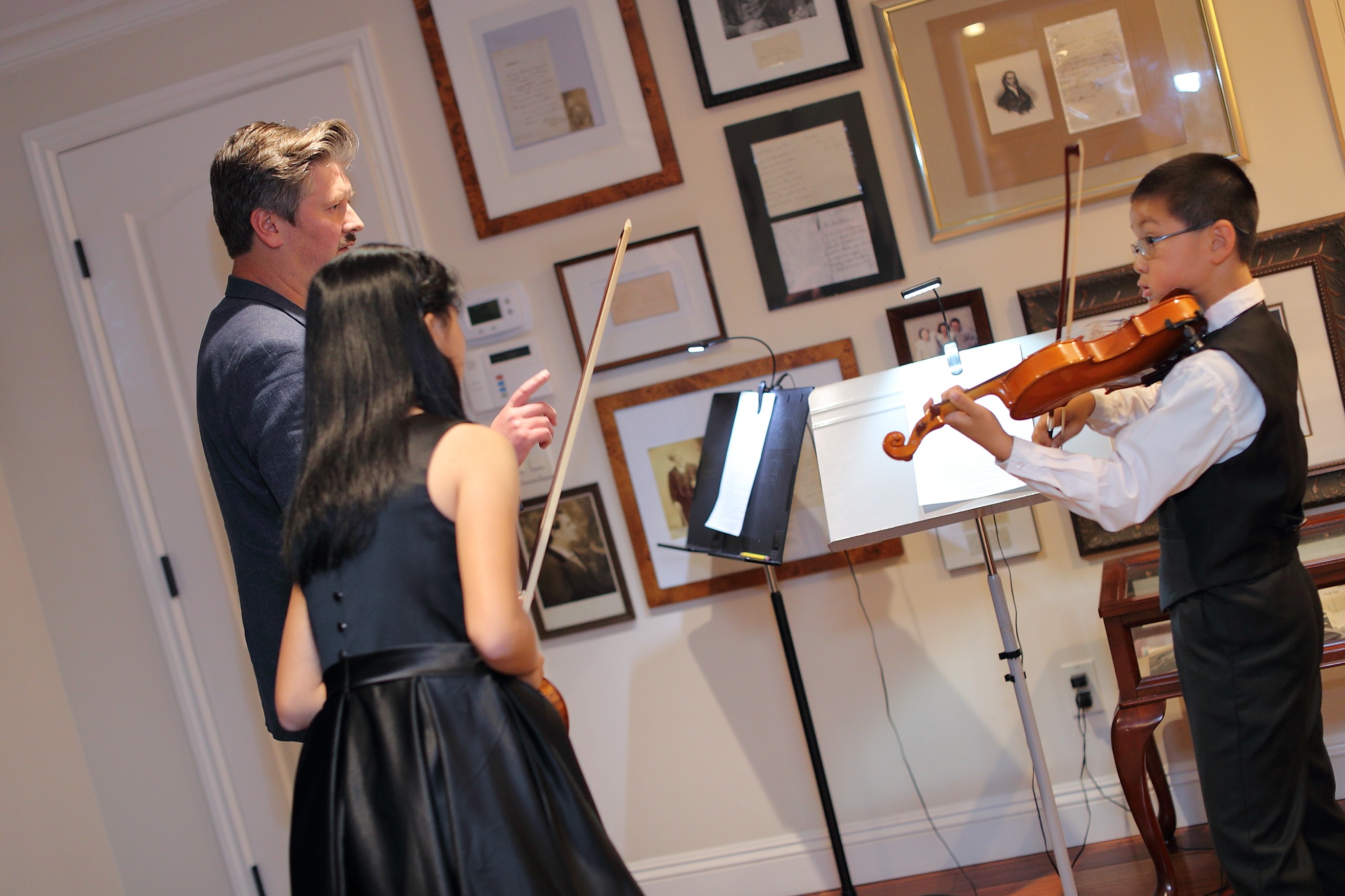 Two children watch a boy playing the violin in an art gallery, with framed artwork on the wall behind them.