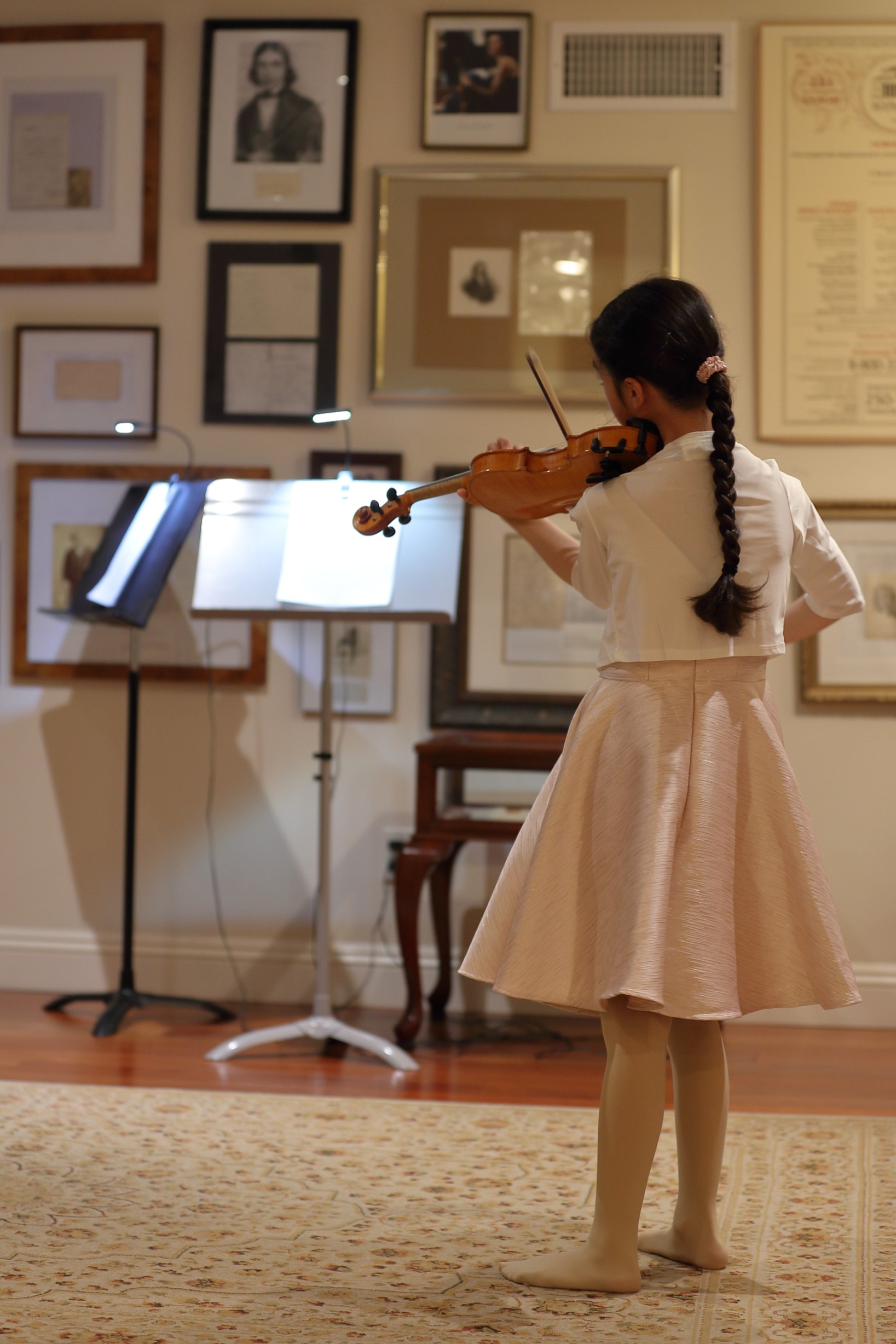 Young girl in a white dress playing violin in a gallery with framed artwork on the wall behind her.