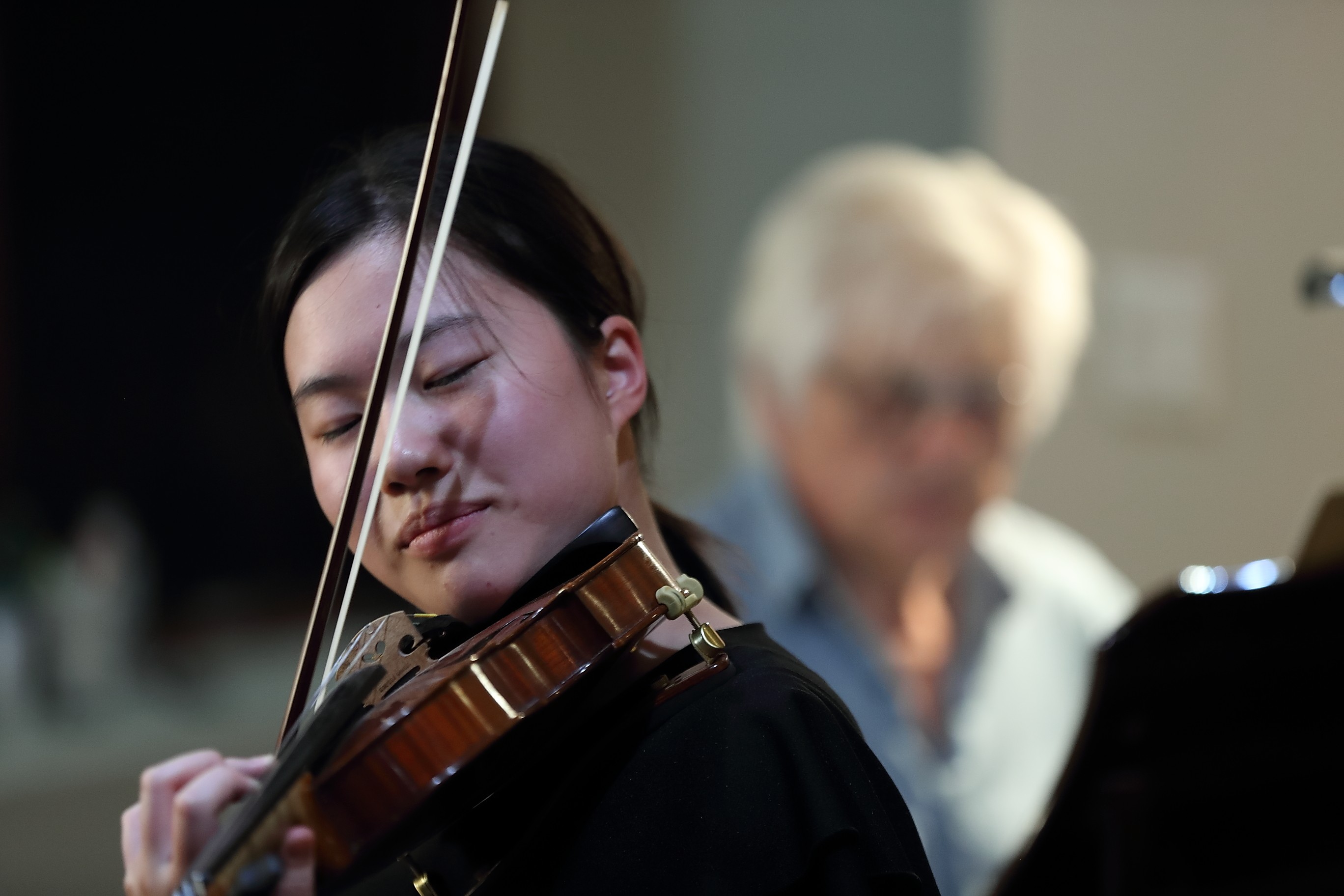 A woman playing the violin with a blurred older woman in the background, both indoors.
