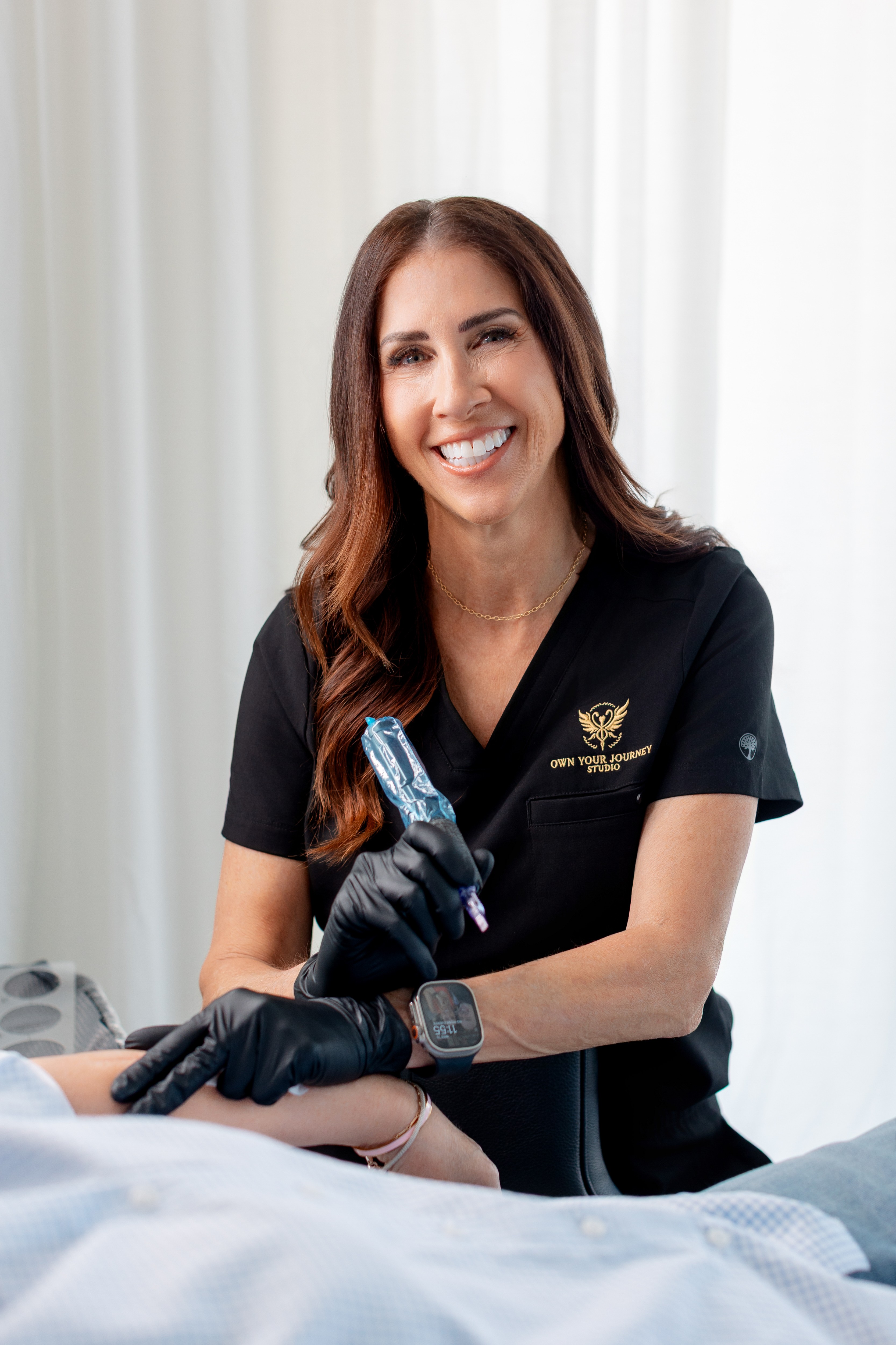 Smiling woman in black scrubs and gloves holding a medical tool, sitting in a clinical setting.