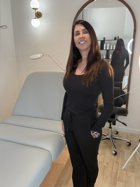 Woman with long dark hair in black outfit taking a mirror selfie in a room with wooden floor and white furniture.