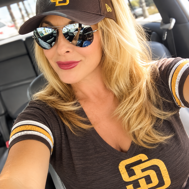 Woman with blonde hair wearing sunglasses, a baseball cap, and a San Diego Padres shirt, sitting in a car.
